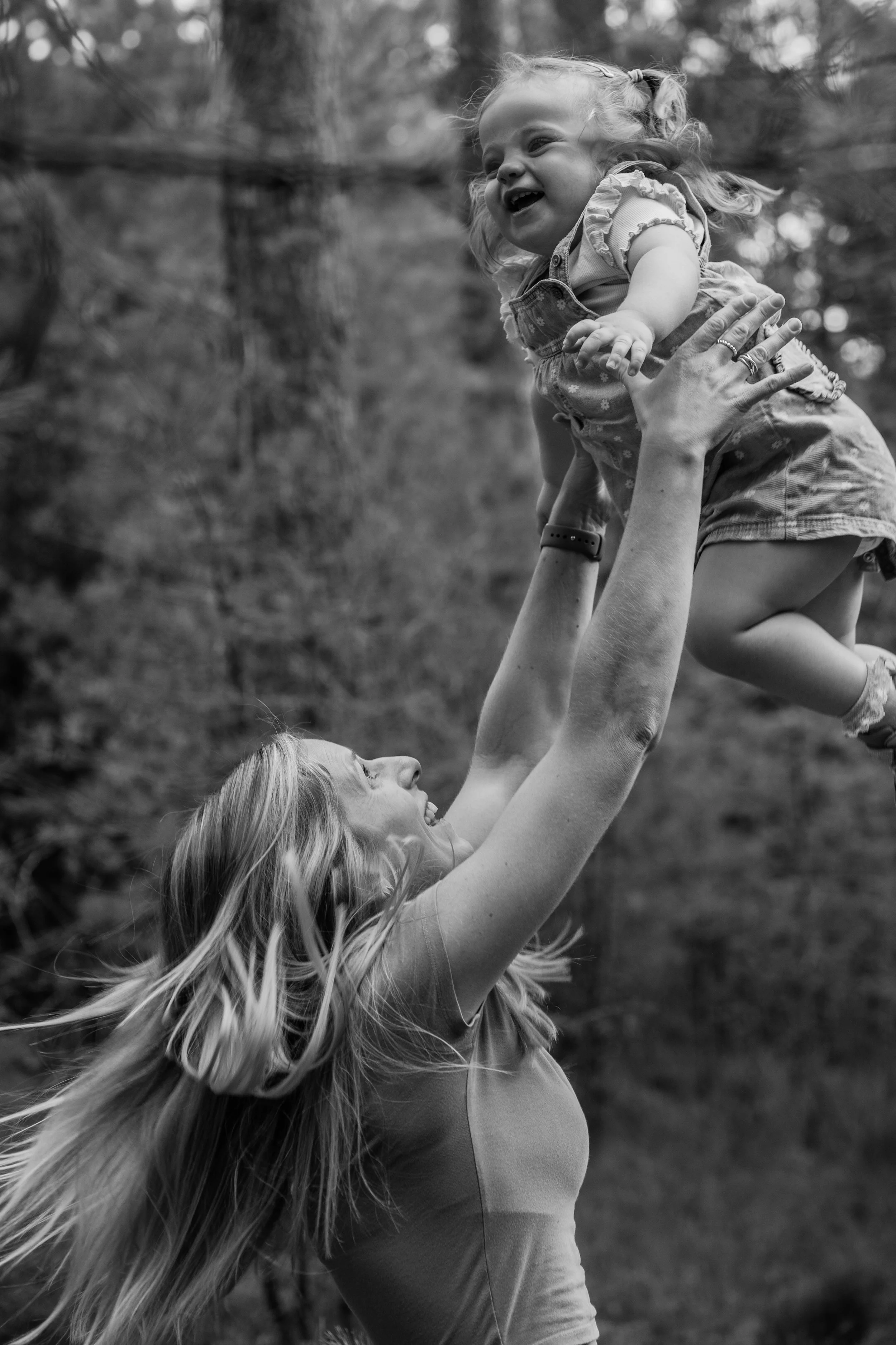 A woman with long hair lifts a young girl in the air outdoors, with trees in the background. The photo is in black and white.
