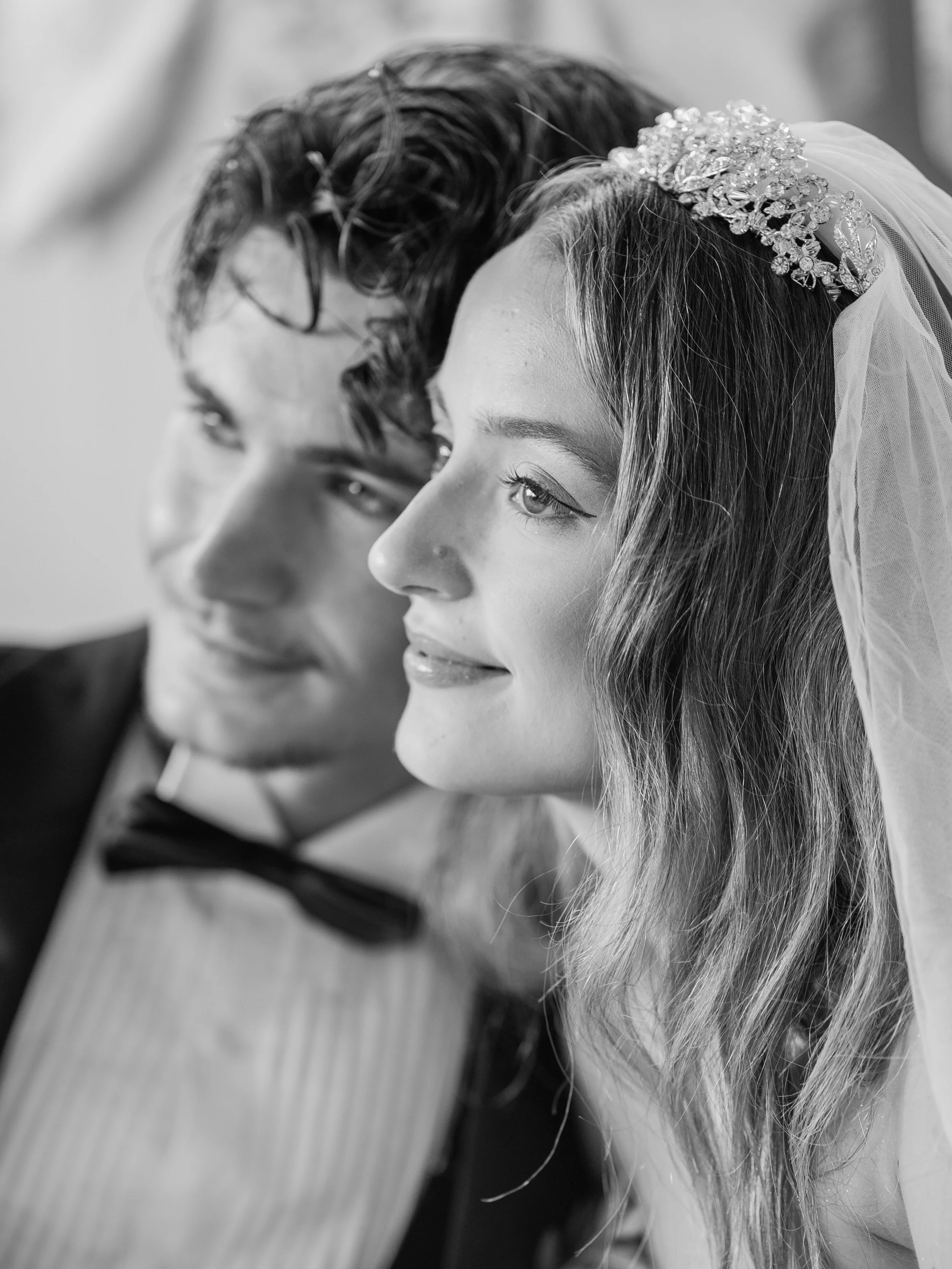 Close-up black and white photo of a bride and groom focusing on their faces, with the bride wearing a jeweled headpiece and veil, and the groom with curly hair.