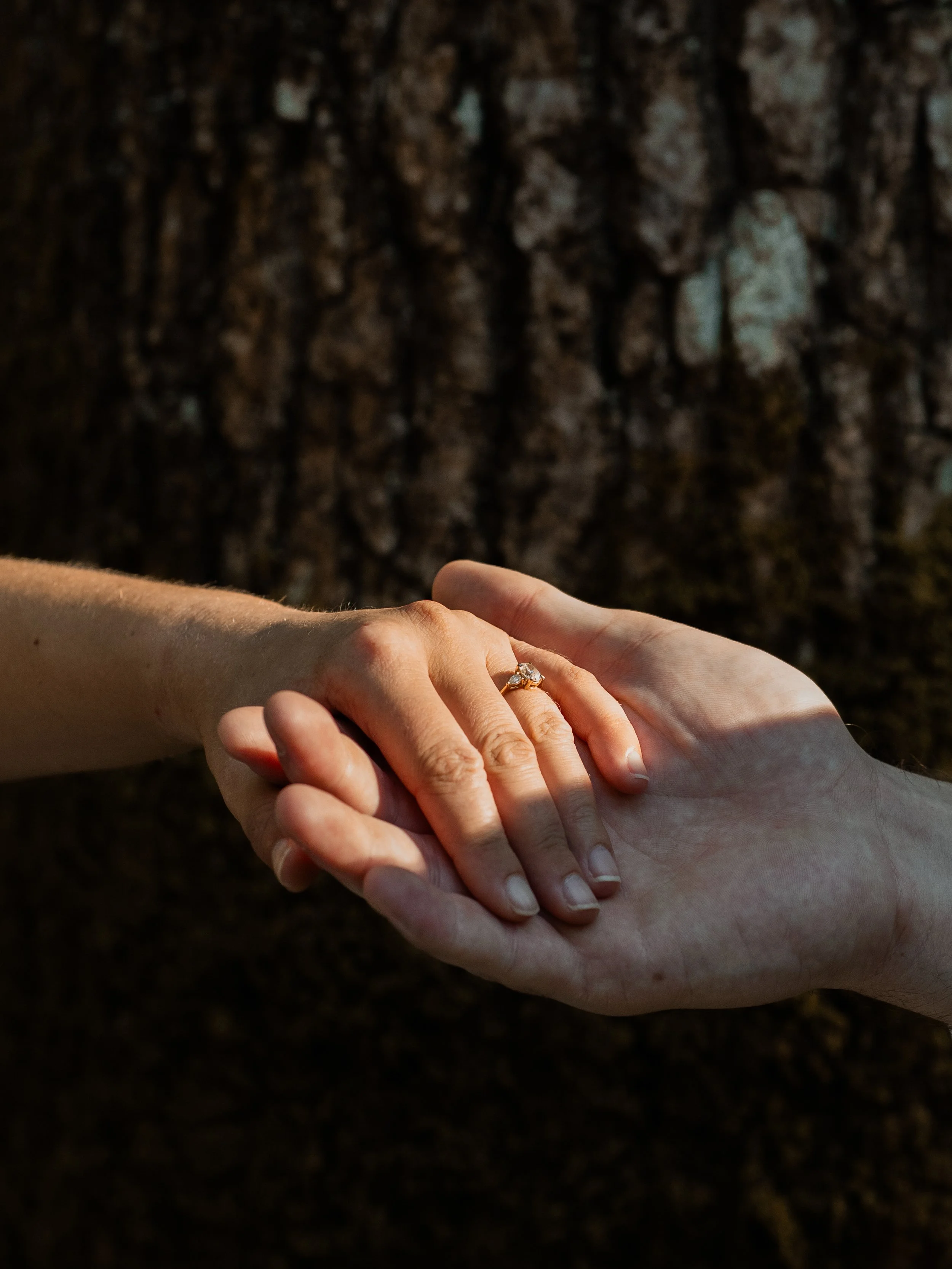 A man and woman holding hands with a visible engagement ring, standing on a textured ground outdoors.