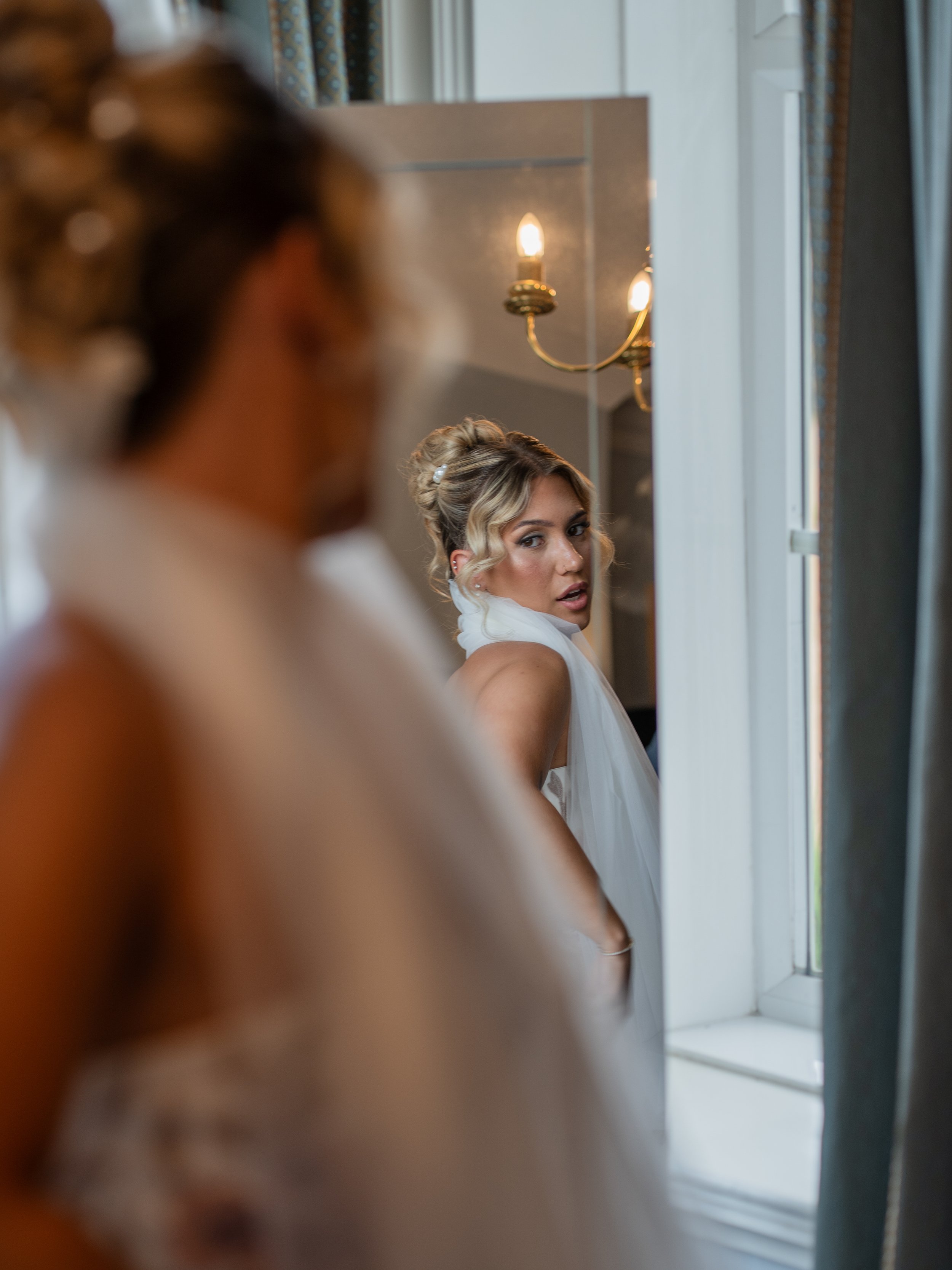 A woman with blonde, curled hair looking into a mirror, dressed in a white gown, in a room with a chandelier and light-colored walls.