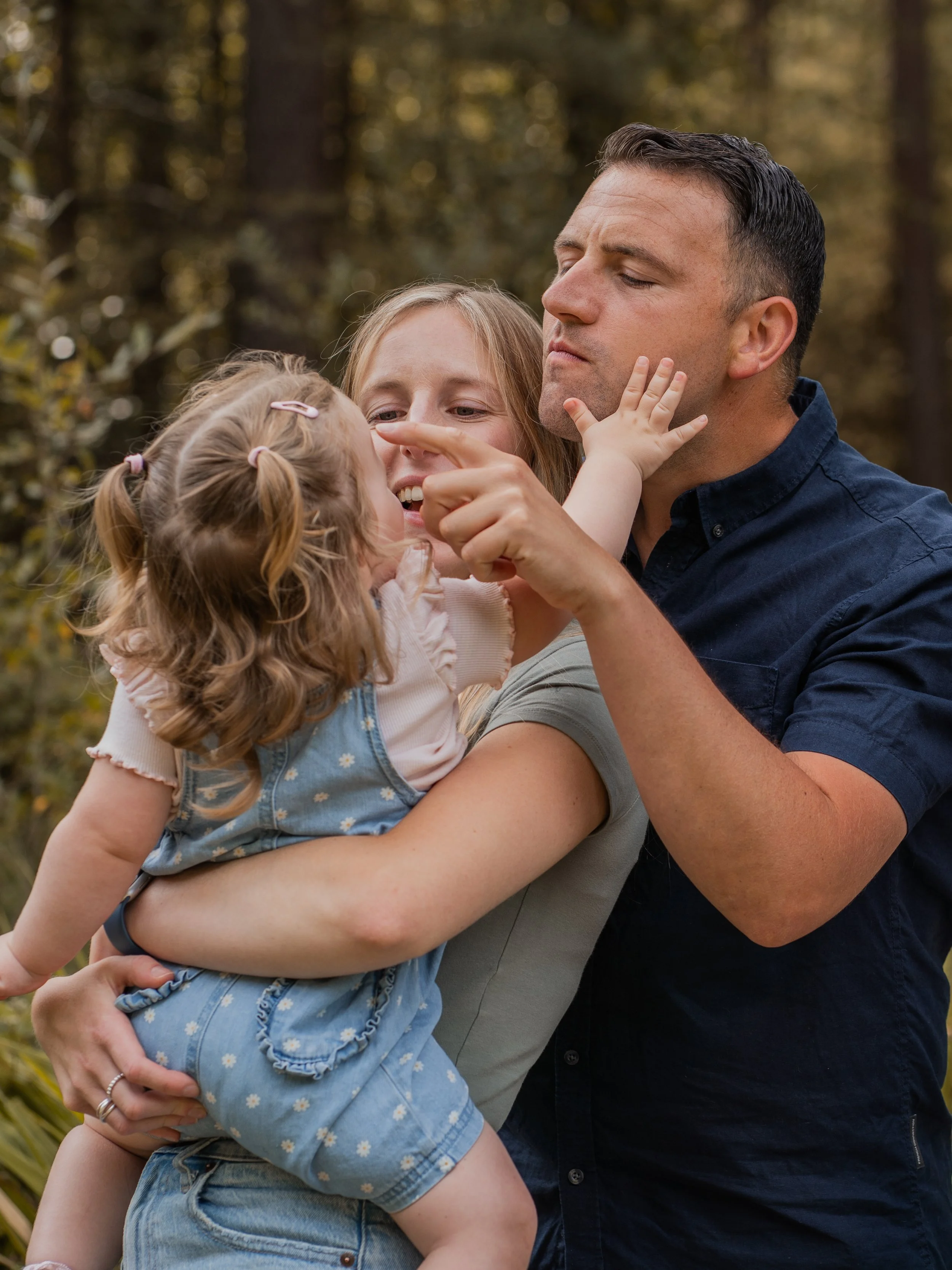 A man holding two young girls in an outdoor setting with trees in the background. The man has short dark hair and is wearing a dark shirt. One girl has curly hair with multiple hair clips, and the other girl has straight hair. The girl with curly hai