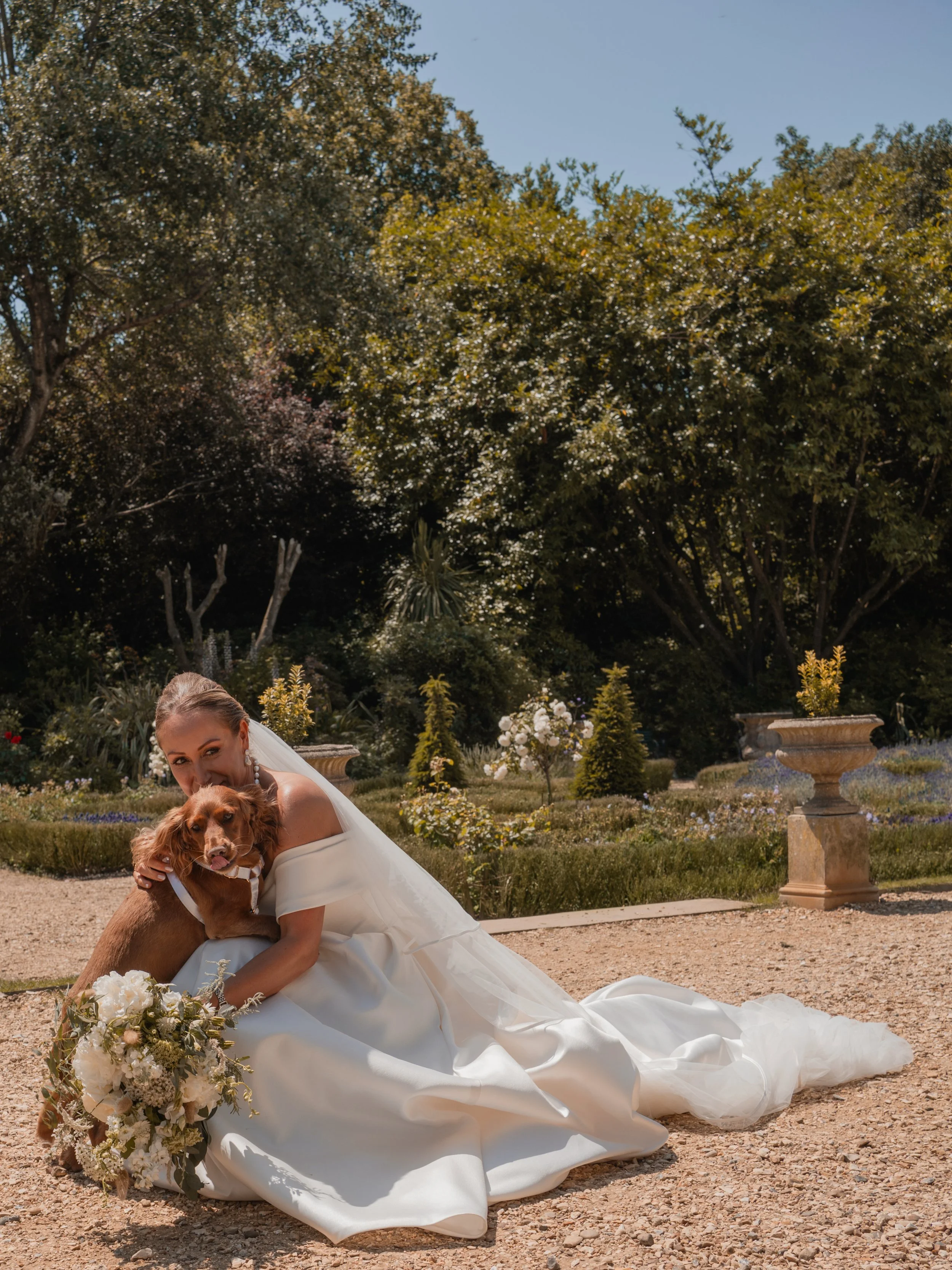 A bride in a white wedding dress drinks from a dog while sitting outside in a garden with trees, bushes, and large decorative planters.
