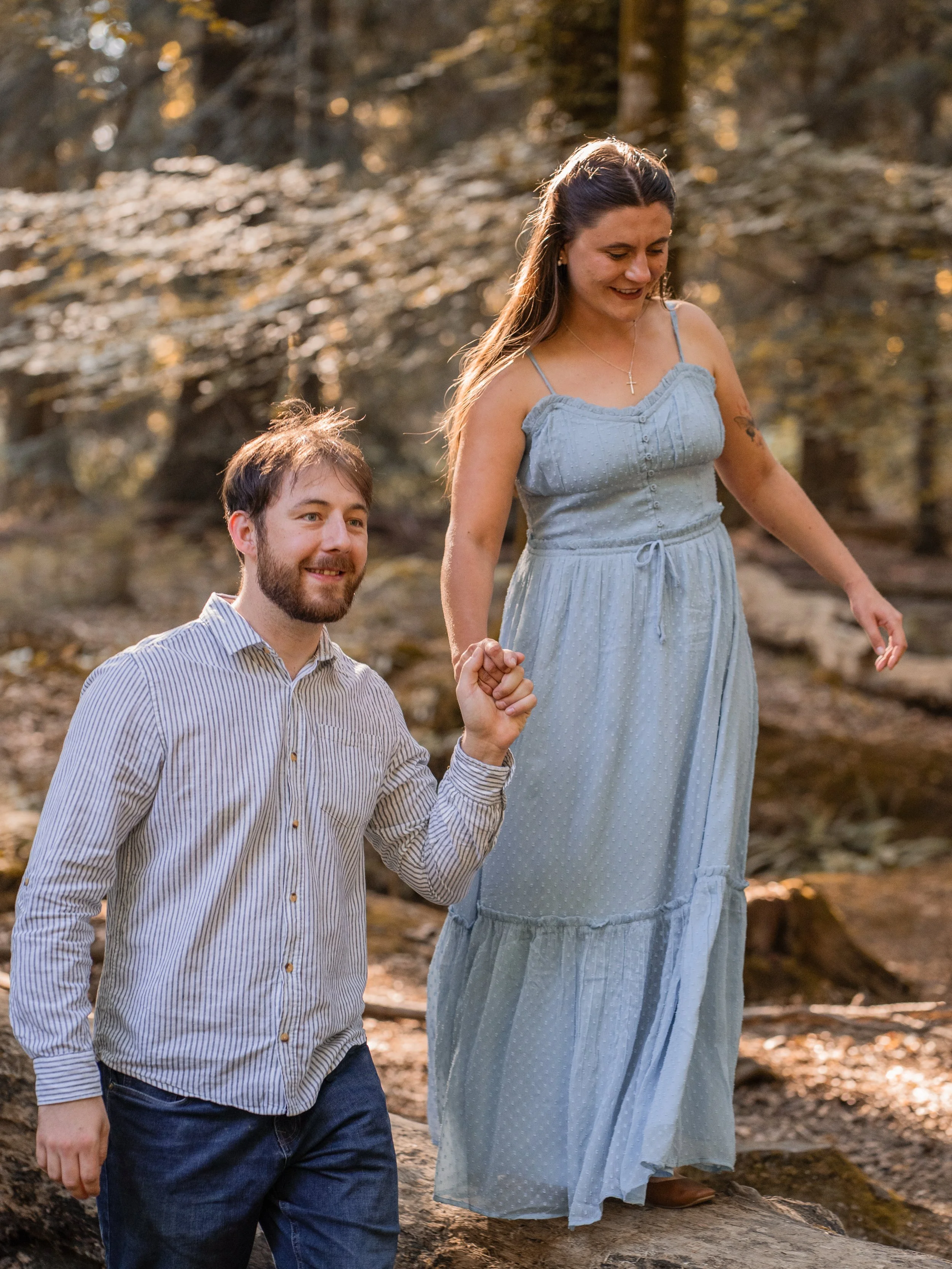 A couple holding hands, walking in a forest with autumn leaves, the woman wearing a light blue dress and the man wearing a striped shirt.