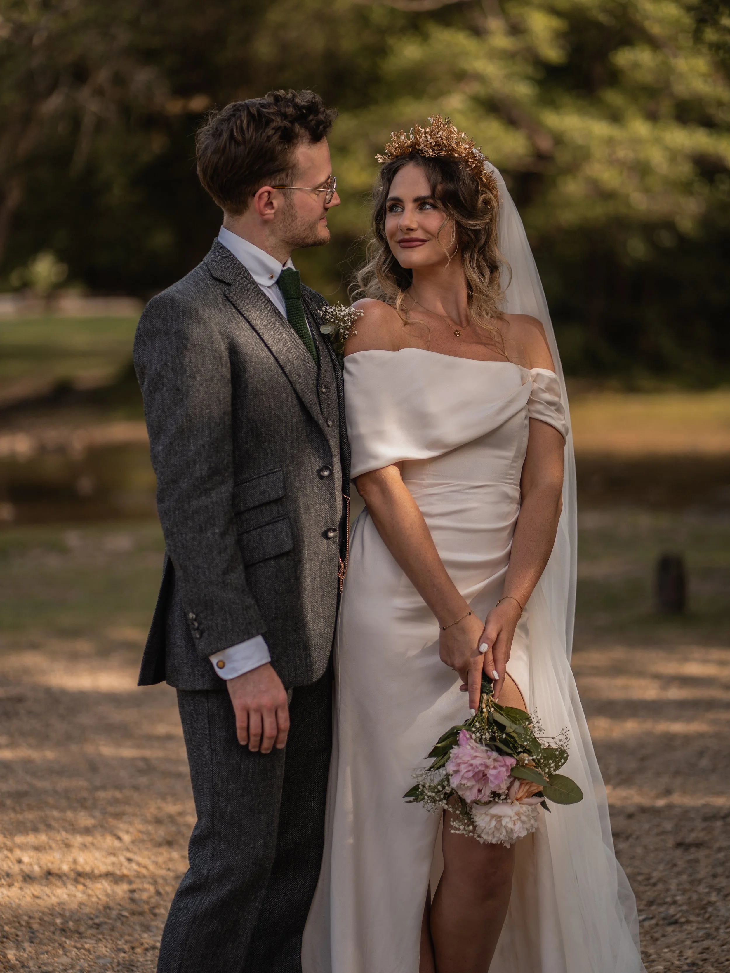 A bride and groom standing outdoors, holding a bouquet of flowers, looking into each other's eyes on a sunny day surrounded by trees.