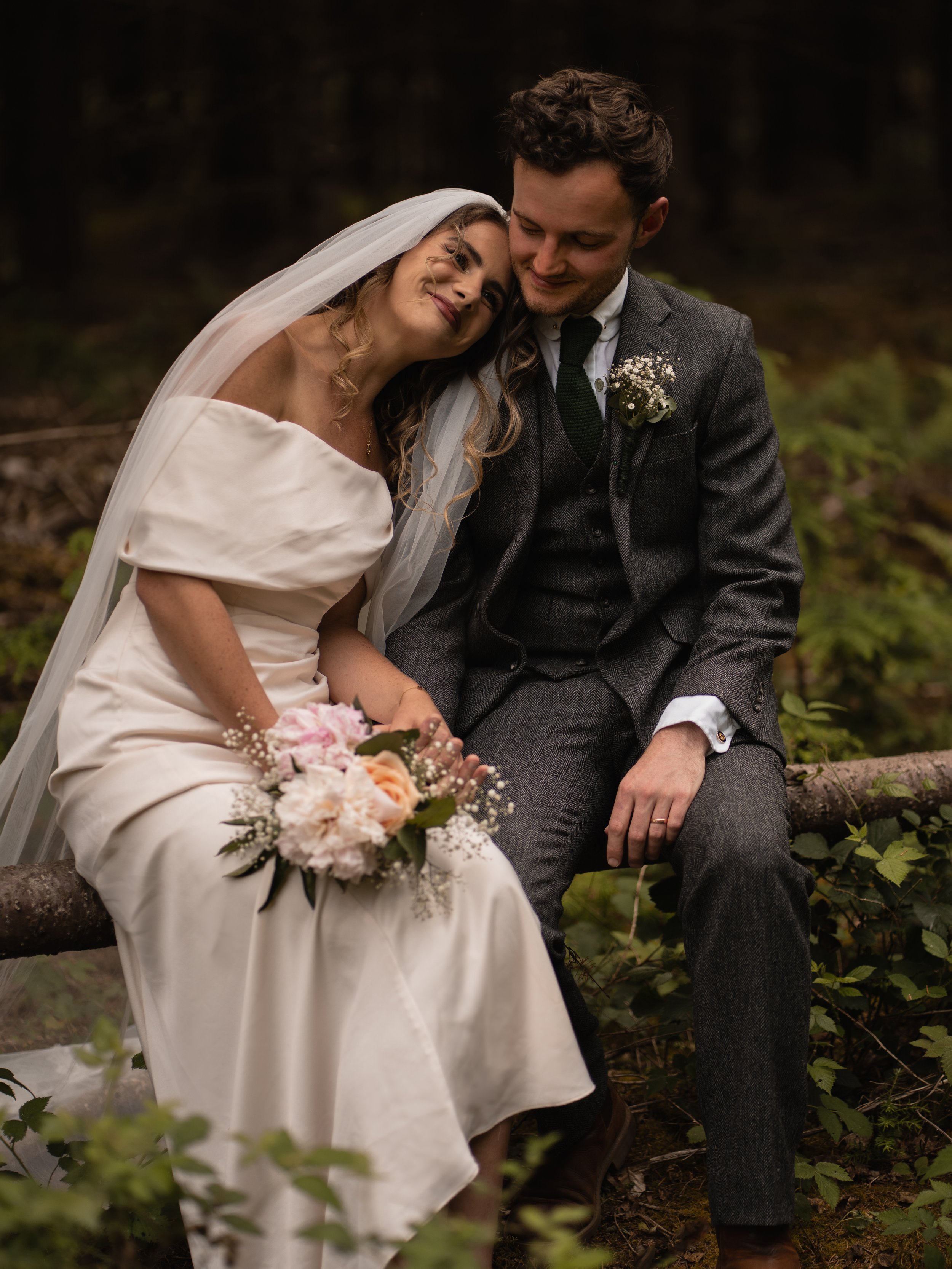 A bride and groom sitting close together outdoors, smiling and embracing in a natural, wooded setting.