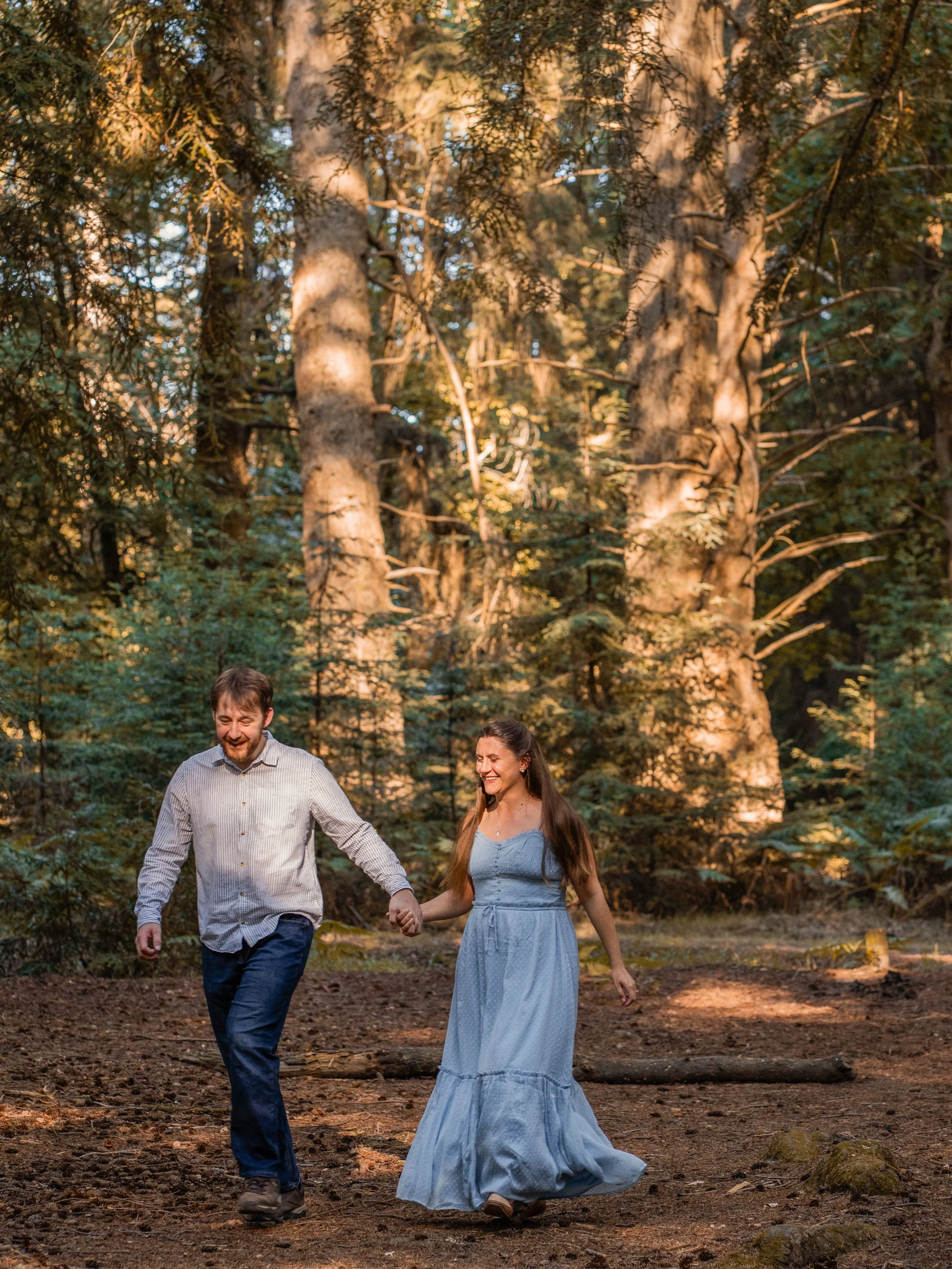 A smiling couple holding hands and walking in a forest during daytime. The man has a beard and is wearing a long-sleeved checkered shirt and jeans. The woman has long hair and is wearing a long, flowing light blue dress.