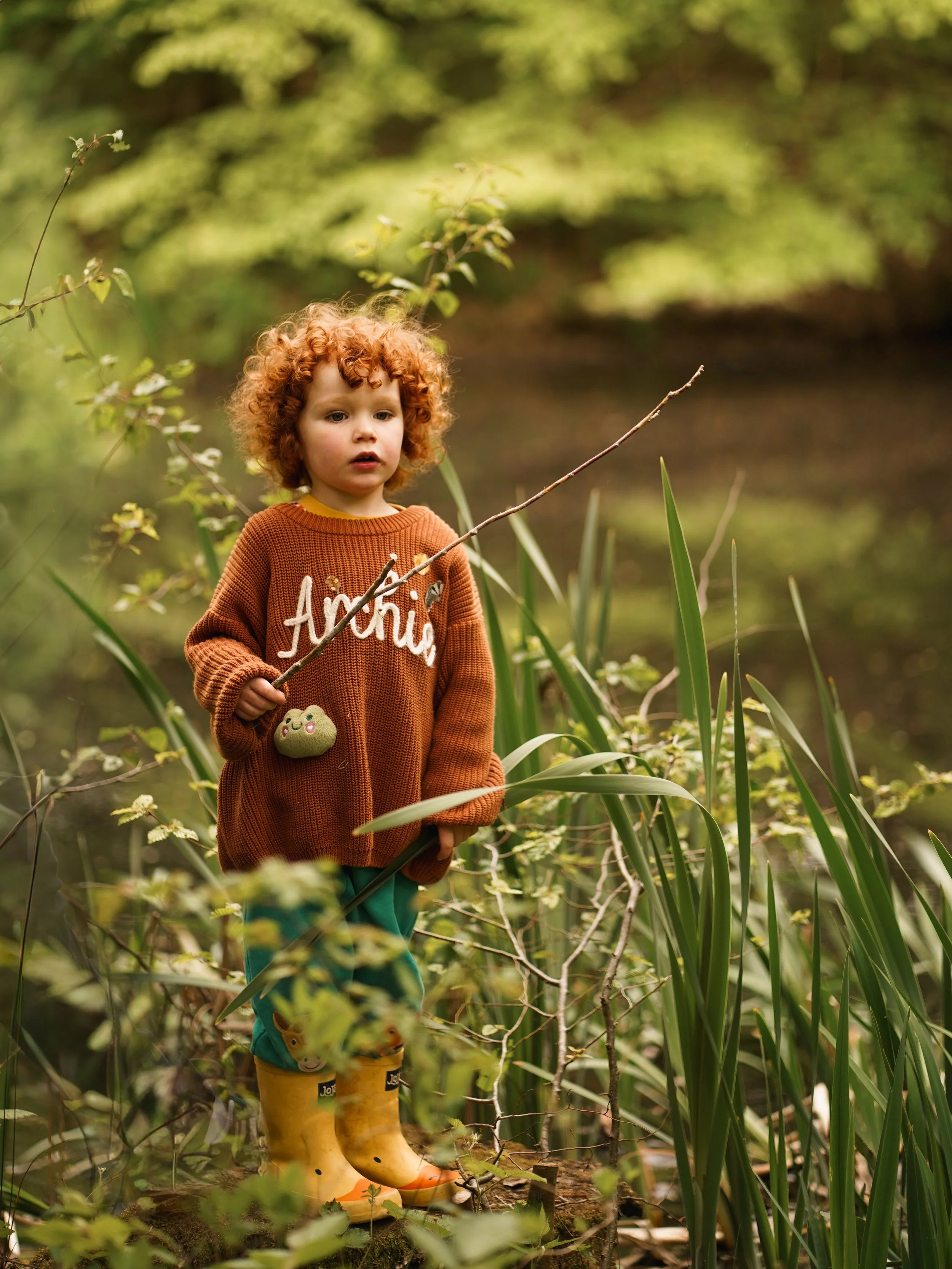 A young curly-haired child standing among tall reeds and plants in a green outdoor setting, holding a stick and wearing yellow rubber boots with a cartoon character design. Editorial family photoshoot in the New Forest