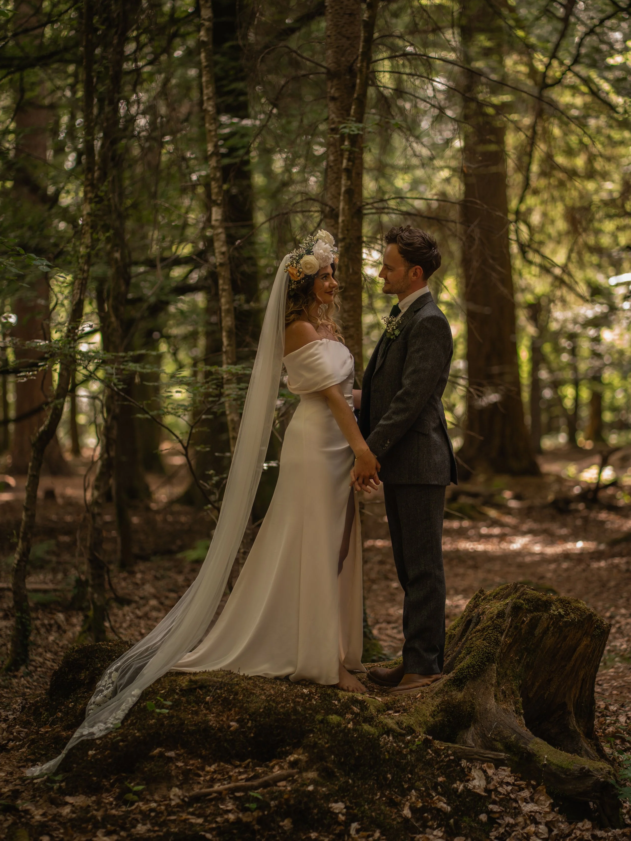 A bride and groom holding hands and looking at each other in a forest. The bride is wearing a white off-shoulder wedding dress with a long veil and a floral headpiece. The groom is dressed in a dark suit with a white shirt. They are standing on a mos