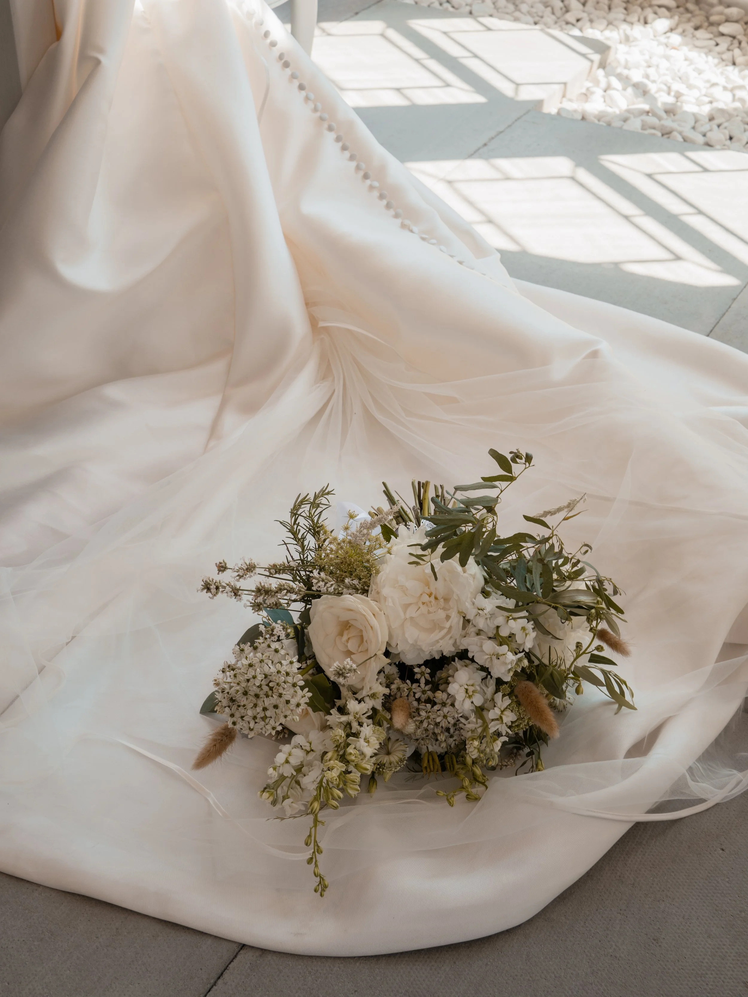Close-up of a white wedding dress with pearl buttons and a bouquet of white flowers and greenery on the fabric.