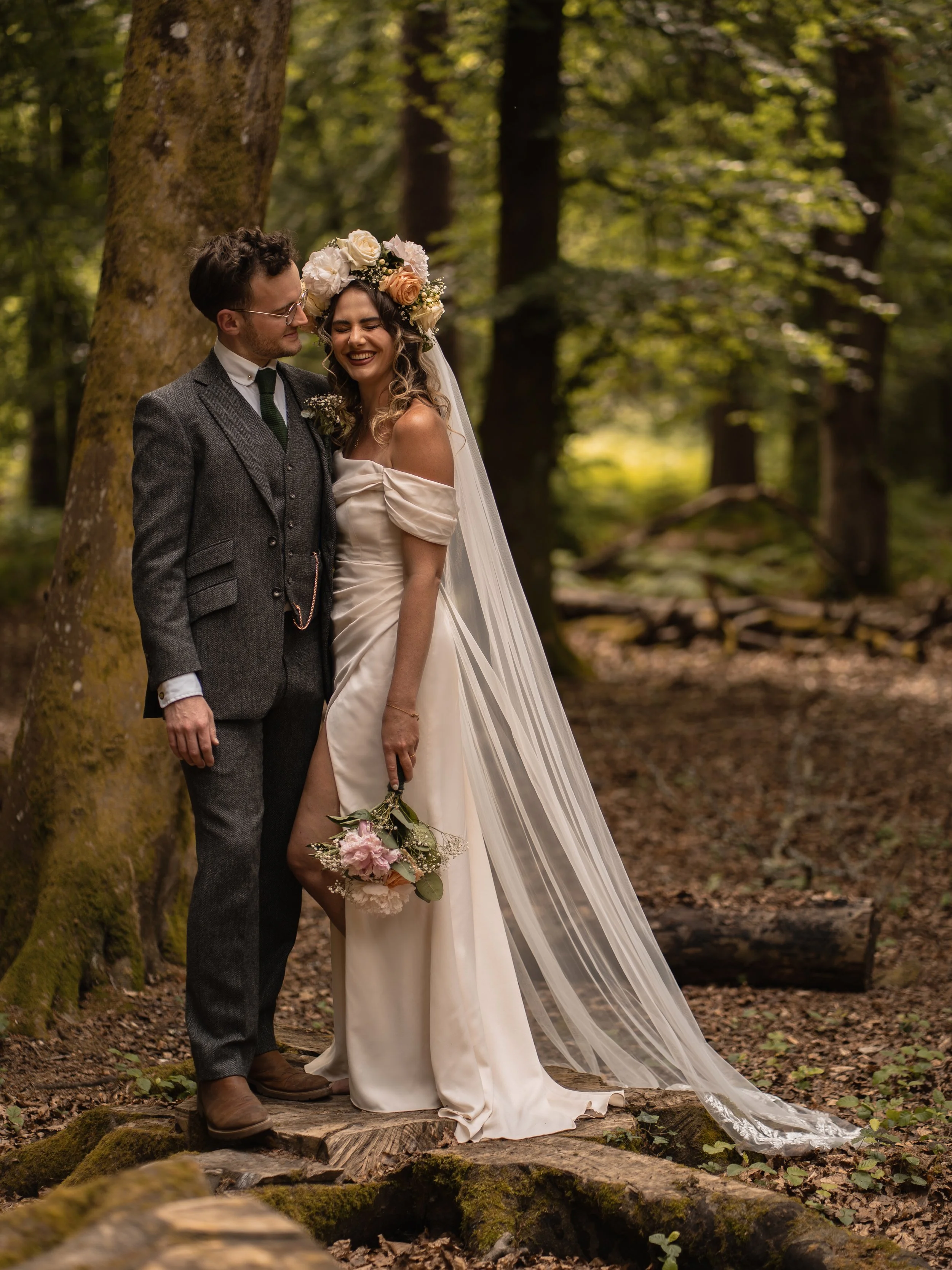 A bride and groom standing together in a forest, smiling and enjoying their wedding day. The bride wears a white off-shoulder dress and a floral crown, holding a bouquet. The groom wears a gray suit with a vest, tie, and glasses. They are close to each other, with trees and fallen leaves around them.