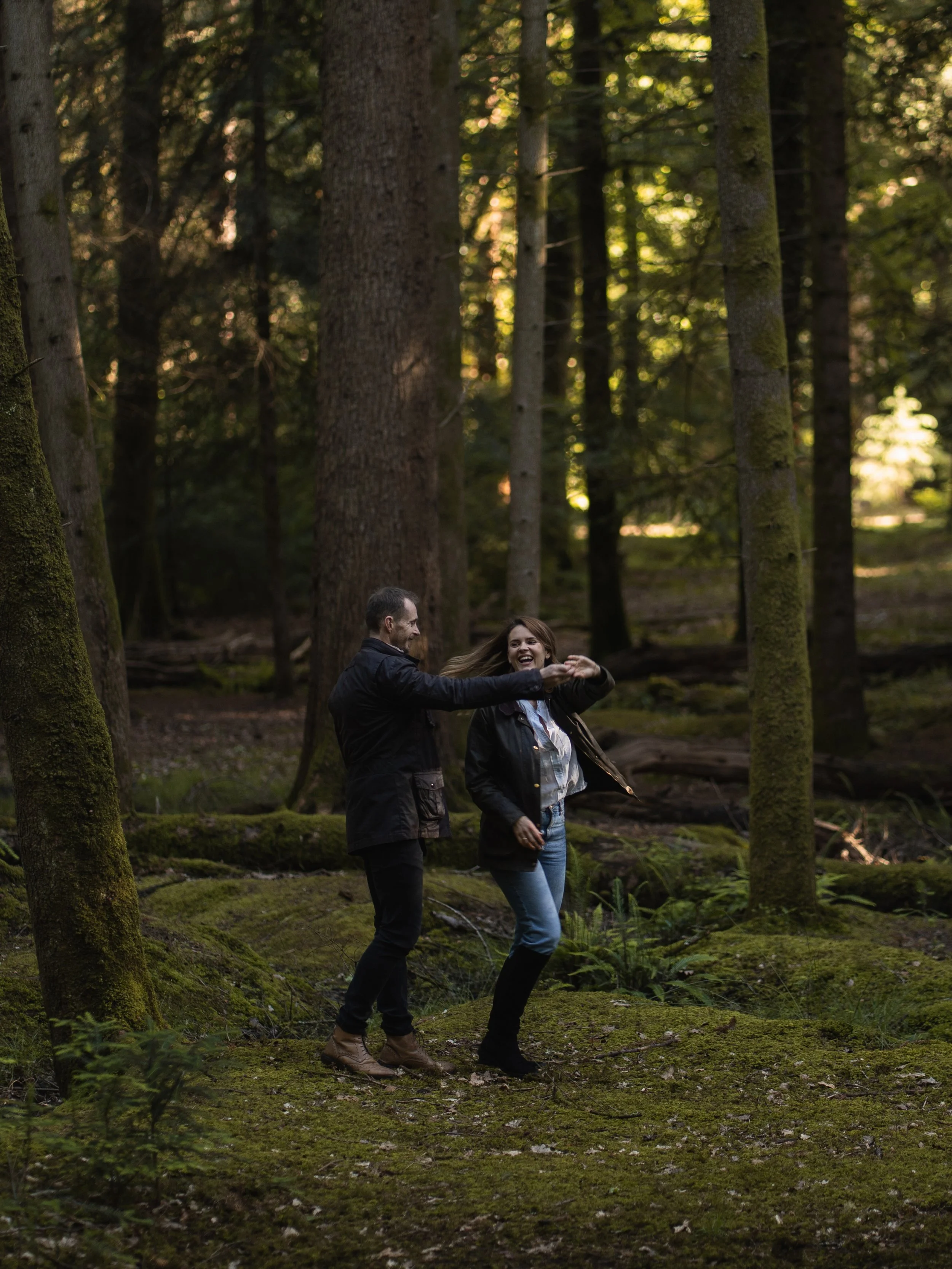 A man and woman are dancing and smiling in a mossy forest surrounded by tall trees and sunlight filtering through the leaves.