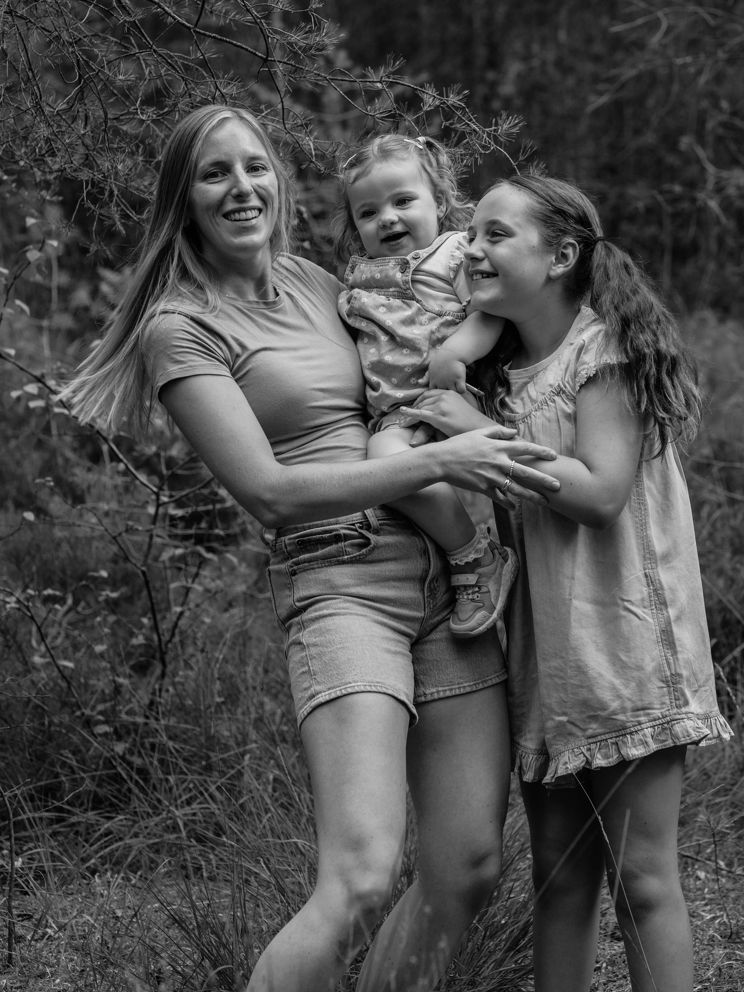 Three women and a young girl standing outdoors in a wooded area. The women are smiling and holding the girl between them. The scene is in black and white.