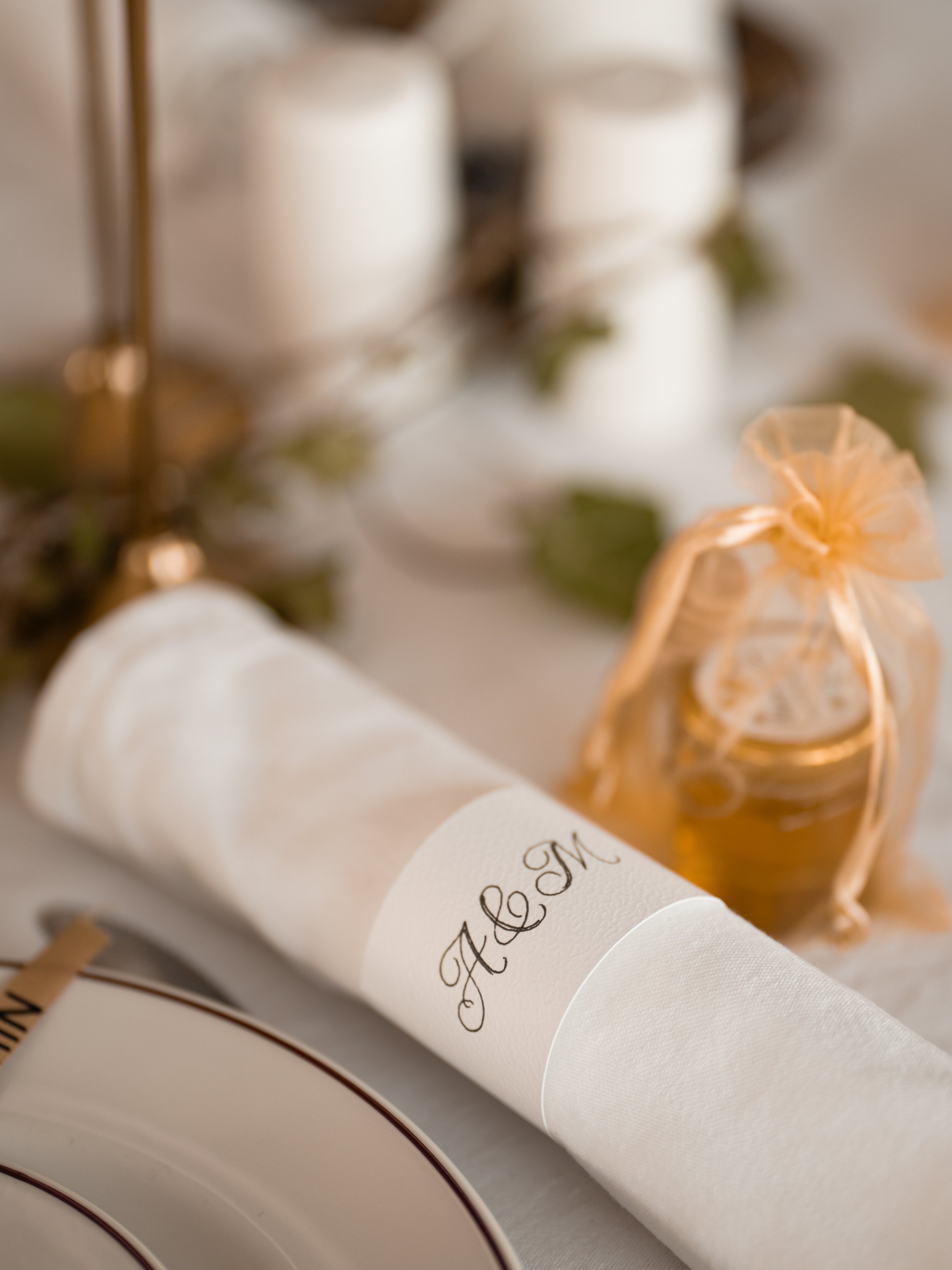 Close-up of a rolled white paper with a handwritten calligraphy message, tied with a gold ribbon, with a blurred background of candles and greenery.