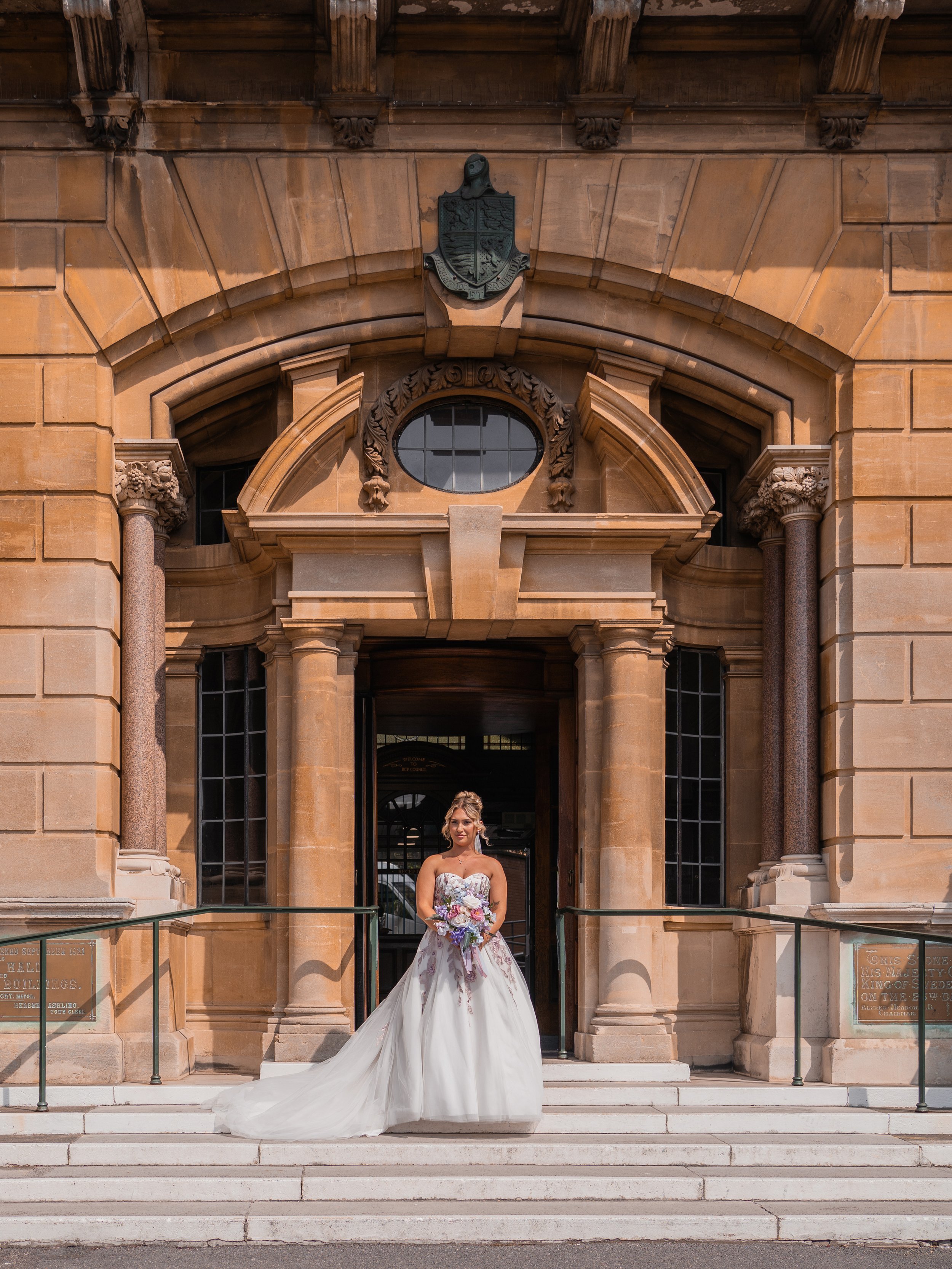 A bride in a strapless wedding gown standing on steps in front of a historic sandstone building with ornate columns and an arched entrance, holding a bouquet of flowers.