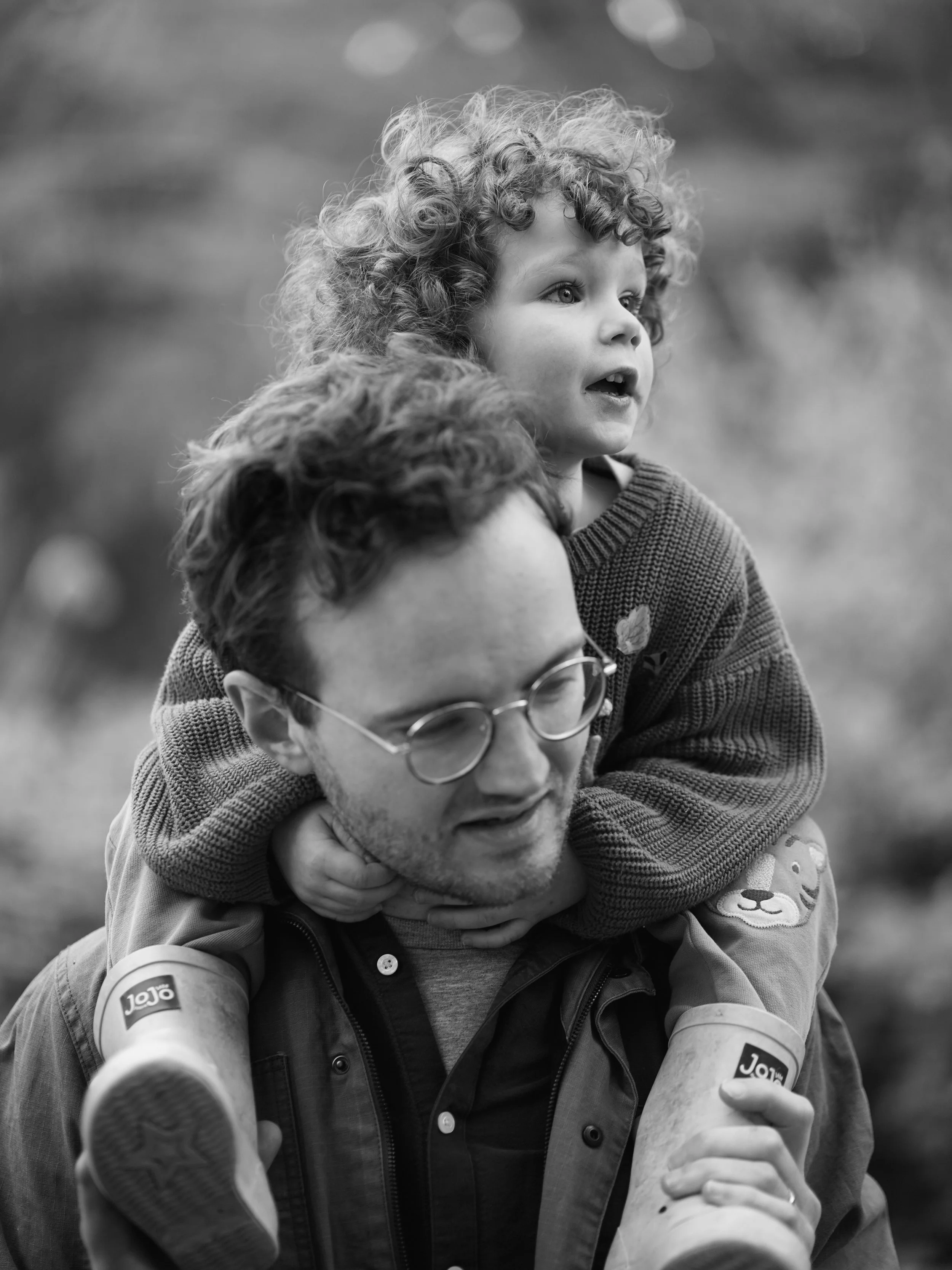 A man with glasses and curly hair gives a piggyback ride to a young girl with curly hair, both smiling and enjoying each other's company outdoors.