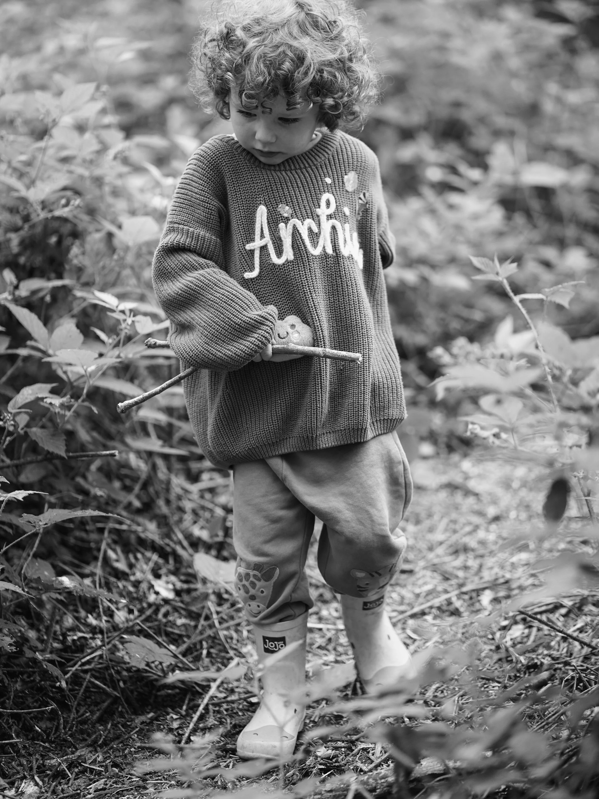 A young child with curly hair wearing a sweater with the word 'Airté' and images of a teddy bear and small bubbles, walking outdoors in leafy surroundings, holding a stick, and wearing rain boots.