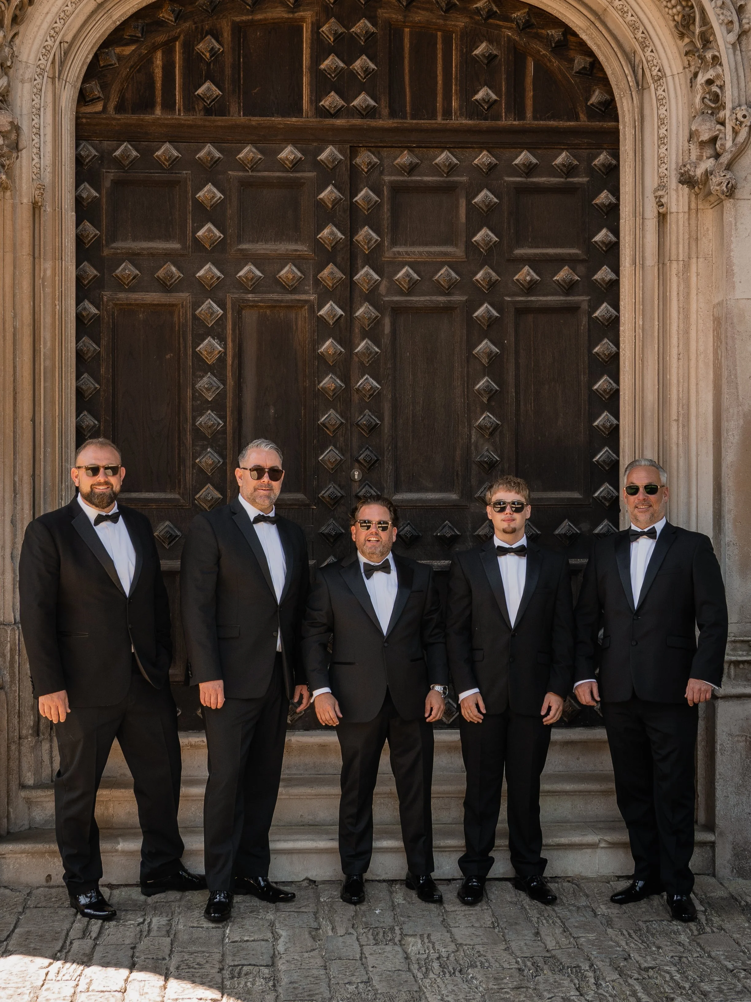 Seven men in black tuxedos with white shirts and bow ties standing in front of a large wooden door with ornate stone framing.
