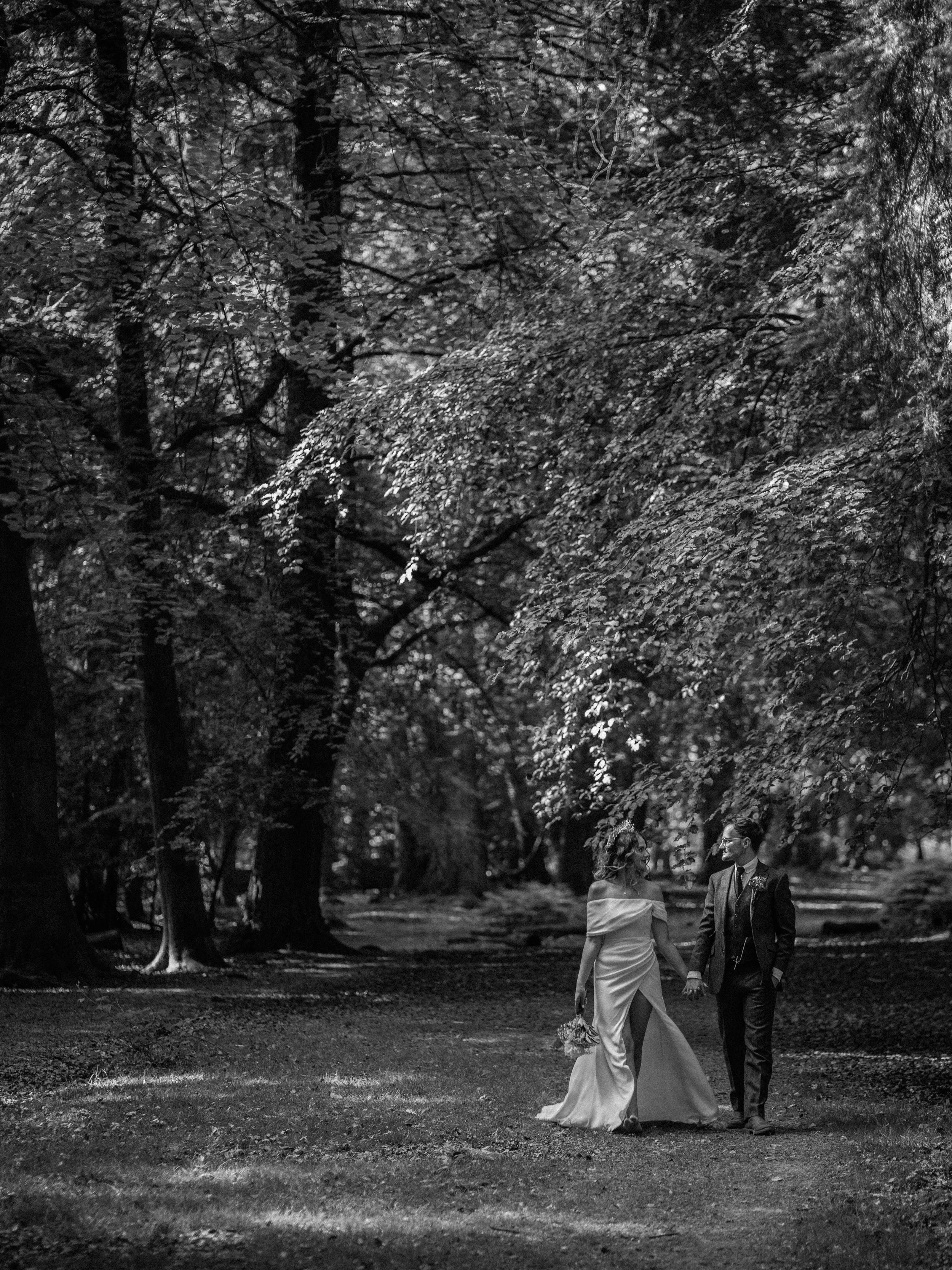 A black and white photo of a bride and groom walking hand in hand in a park surrounded by large trees.