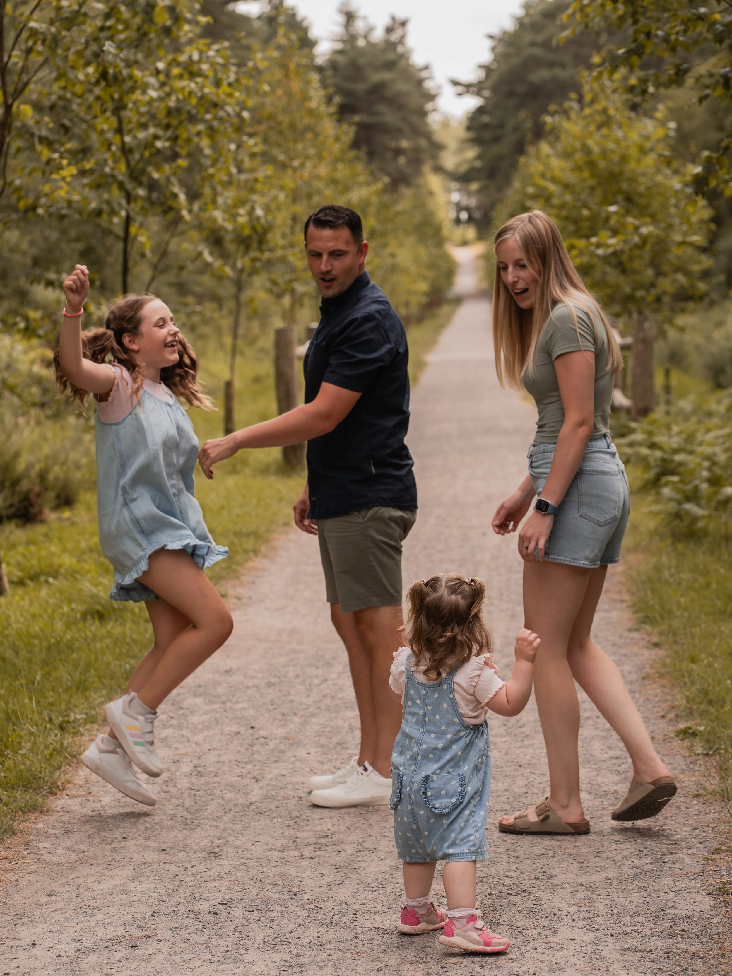 Family of five children and two adults playing and jumping in a park on a gravel path, surrounded by trees with green leaves in summer.