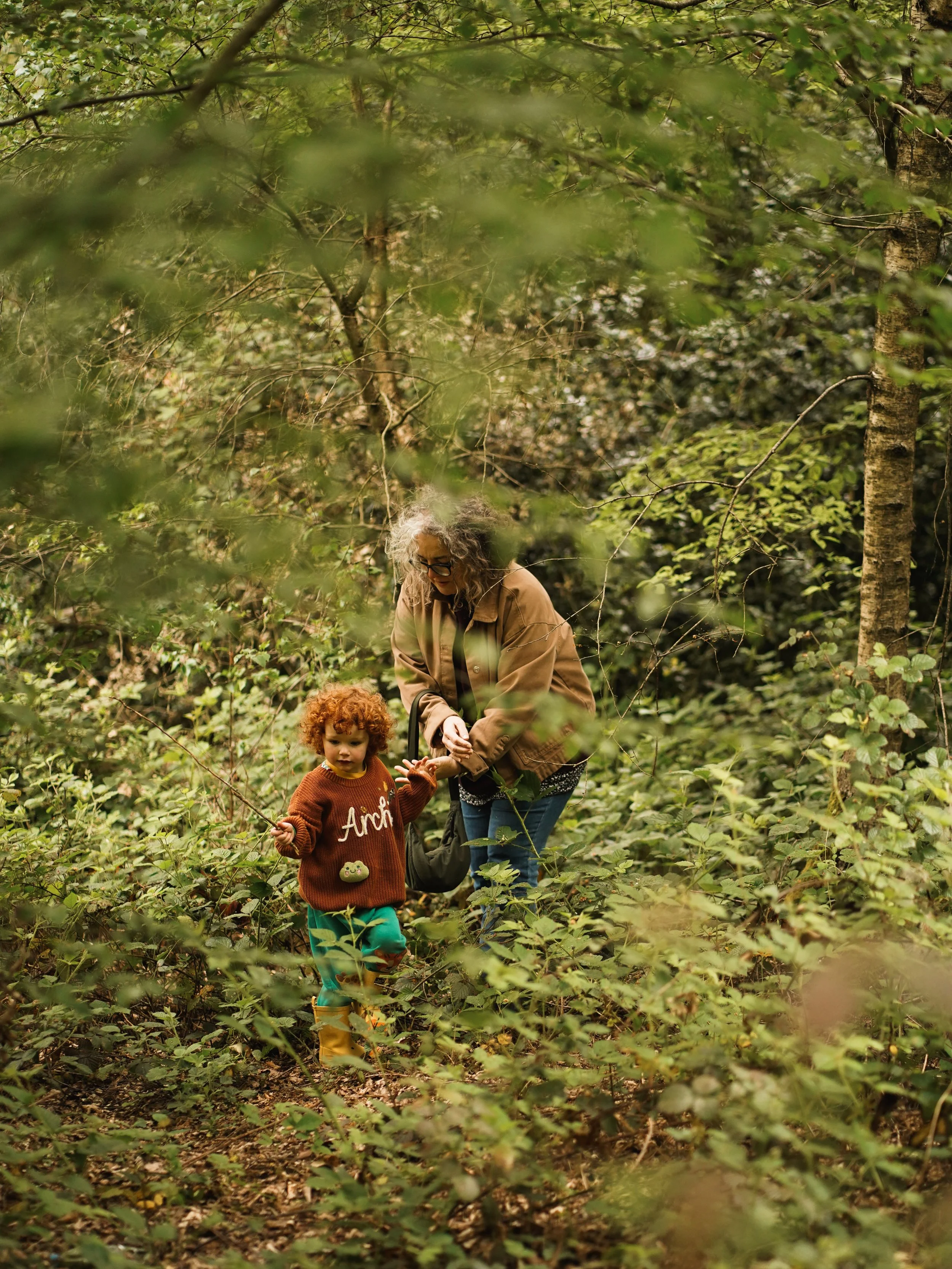 A woman and a young girl exploring a dense green forest, surrounded by leafy plants and trees, with the woman assisting the girl as they walk through the woods.