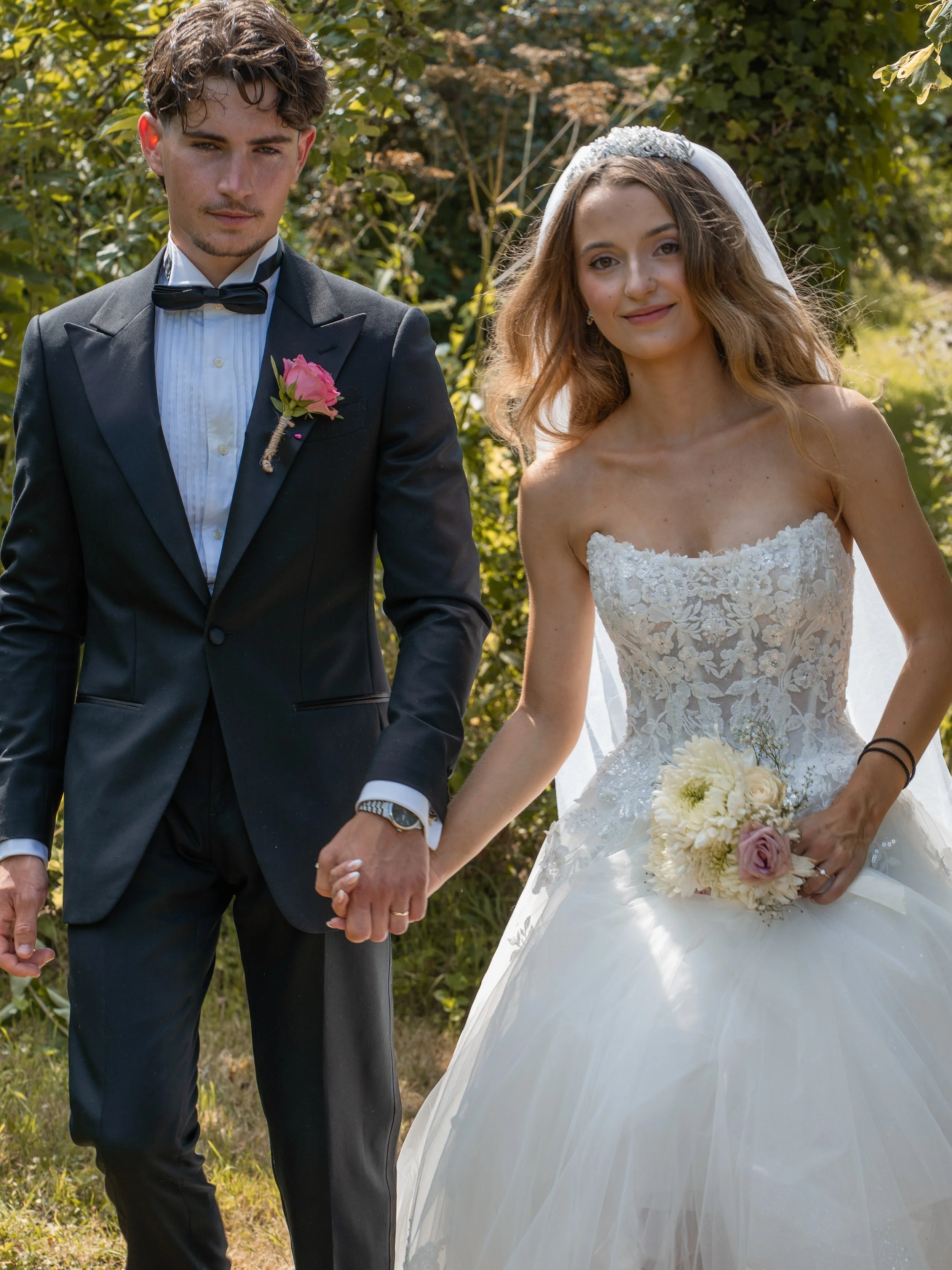 A newlywed couple walking outdoors holding hands, the groom wears a black tuxedo with a pink boutonniere, and the bride wears a white wedding dress with lace details and a veil, holding a bouquet of cream and pink flowers.