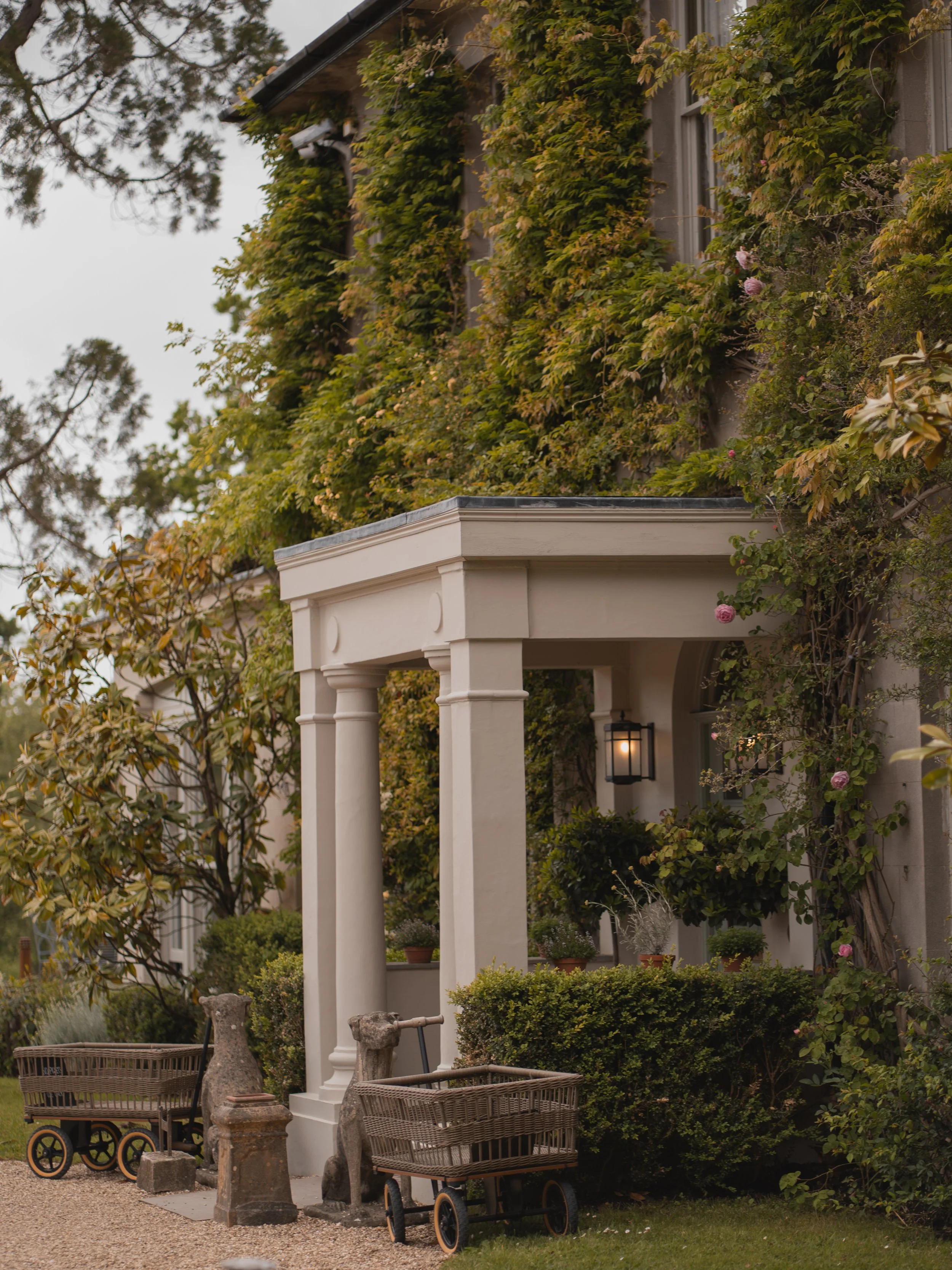 A house with ivy growing on the exterior wall, a porch with classic columns, surrounded by trees, garden furniture, and decorative stone animals, with a lit outdoor lantern near the door.