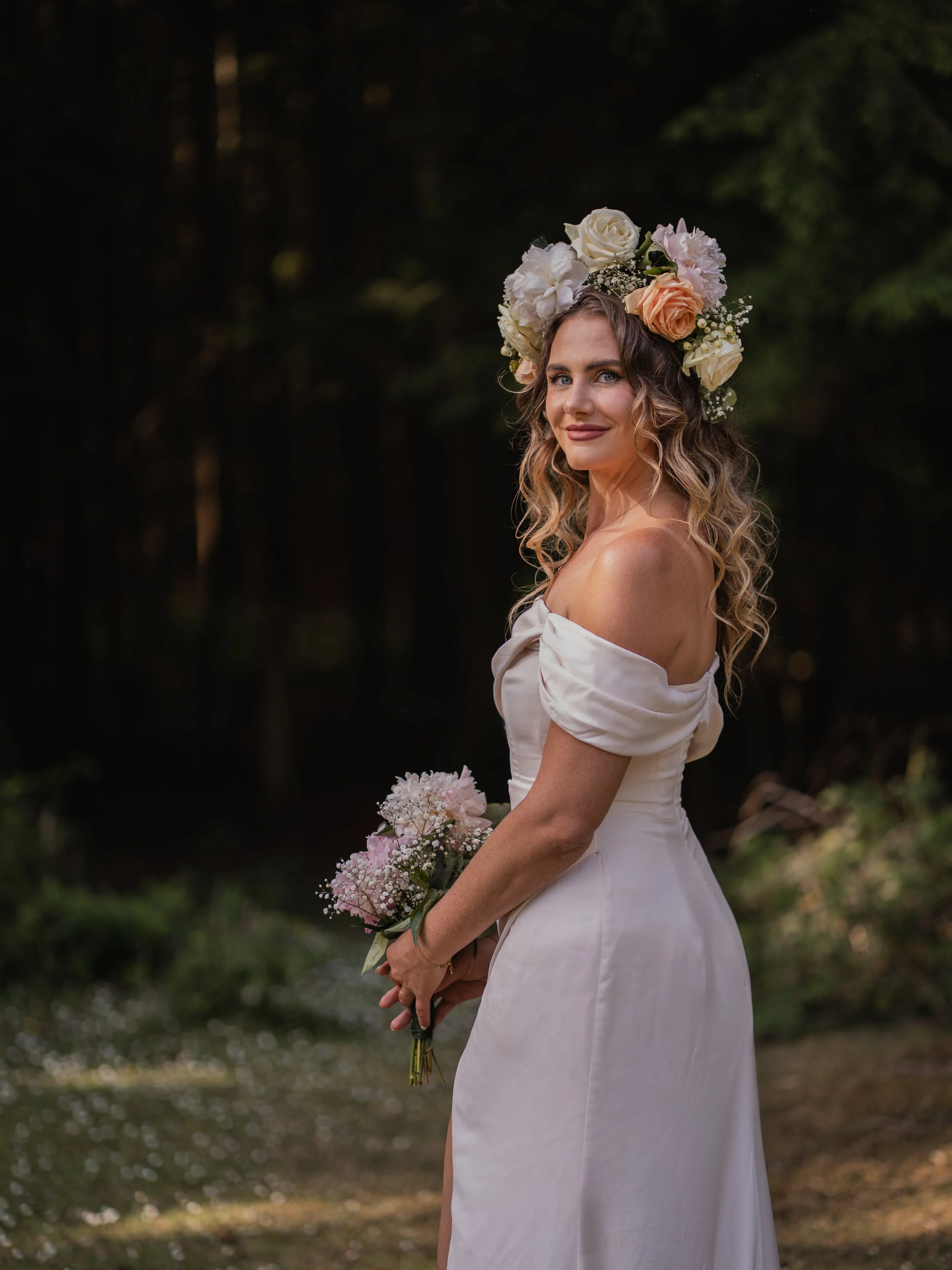 A woman in a white off-the-shoulder dress with a floral crown of white, peach, and pink flowers, holding a bouquet of pink and white flowers, in a dark outdoor setting.