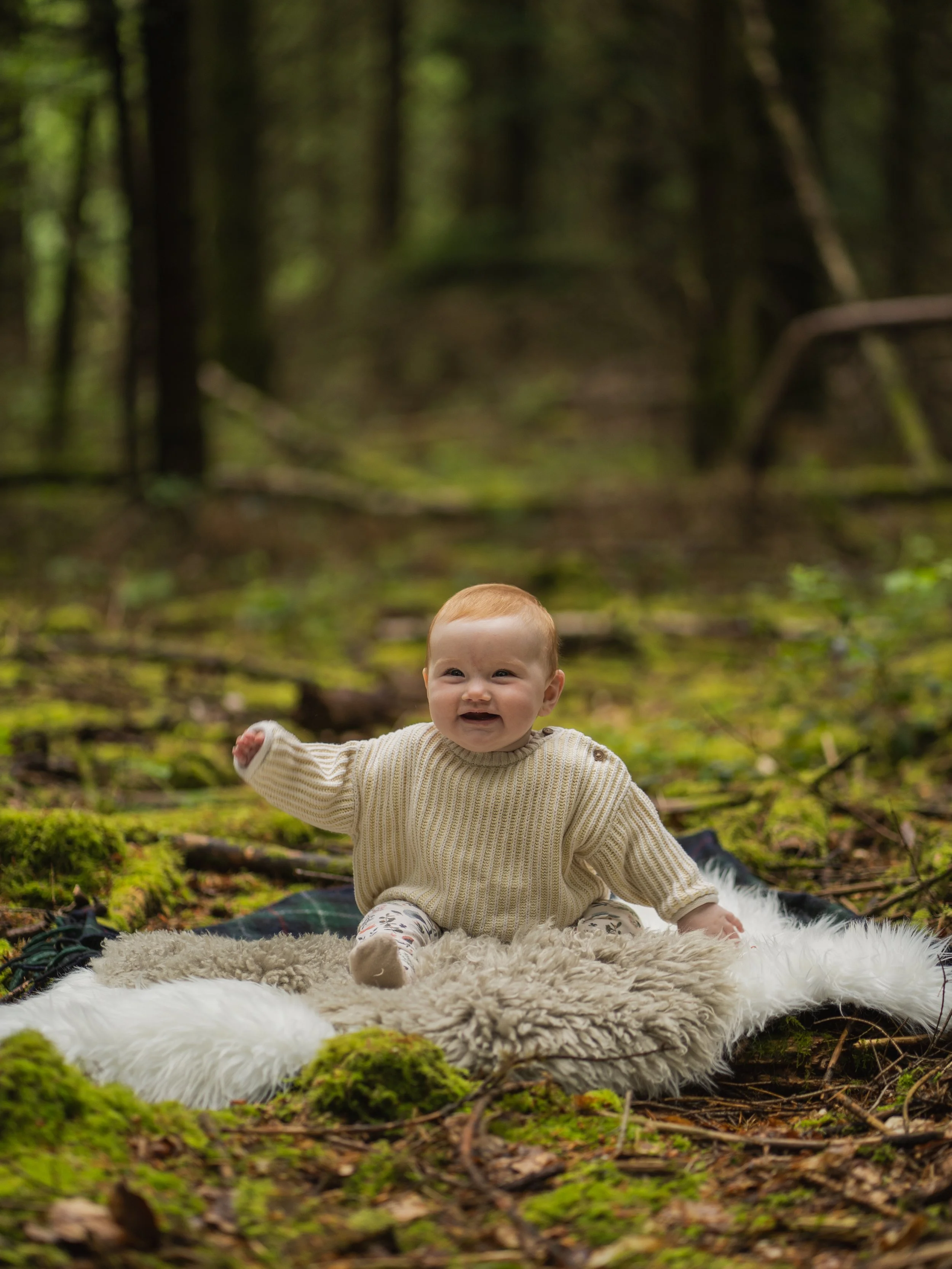 A smiling baby with red hair sitting on a fluffy rug in the woods surrounded by green moss and trees in Wilverly in Brockenhurst in the New Forest for a family photoshoot.