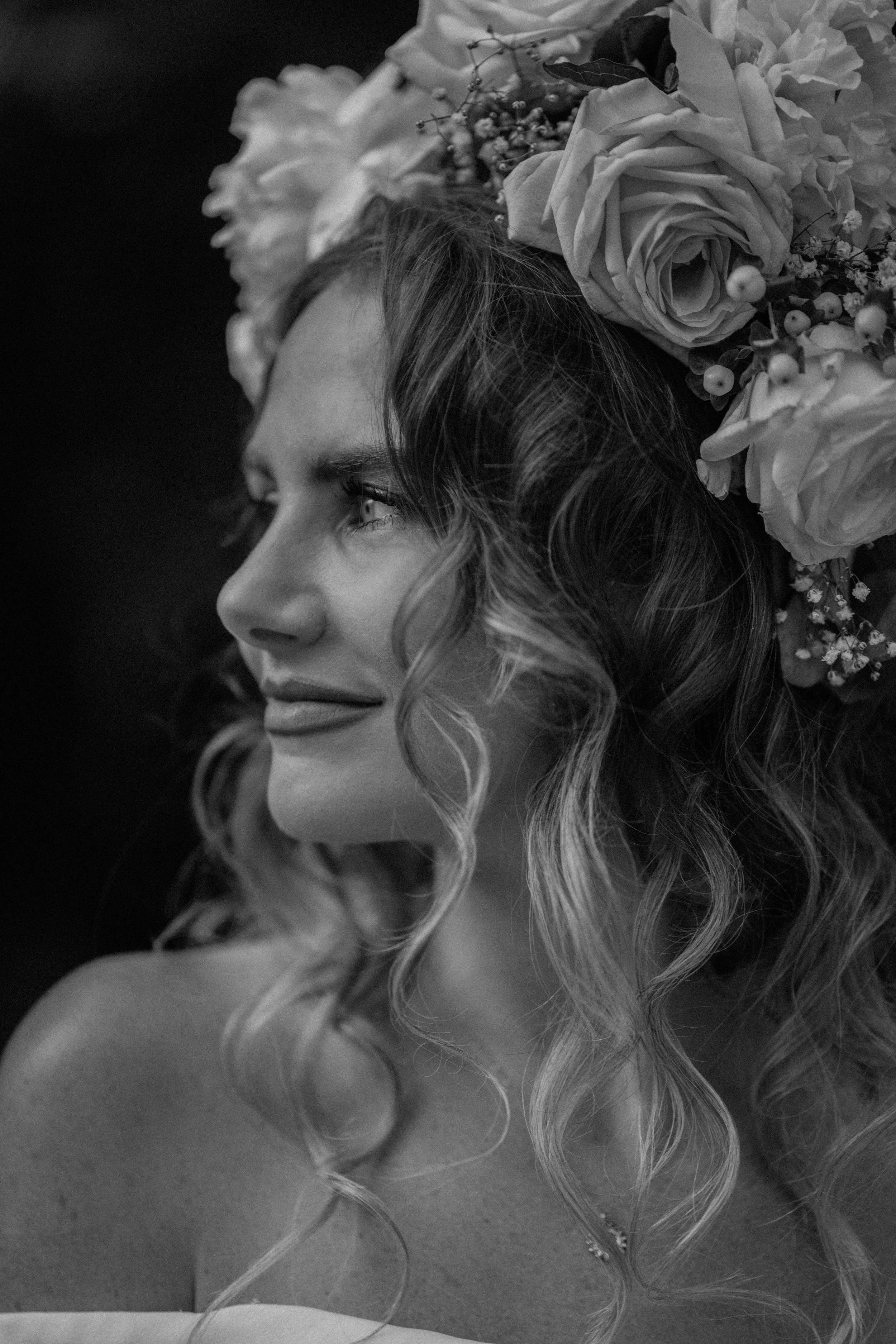 A black and white close-up of a young woman with long, wavy hair adorned with a large floral headpiece, featuring roses and small berries, and looking off to the side.