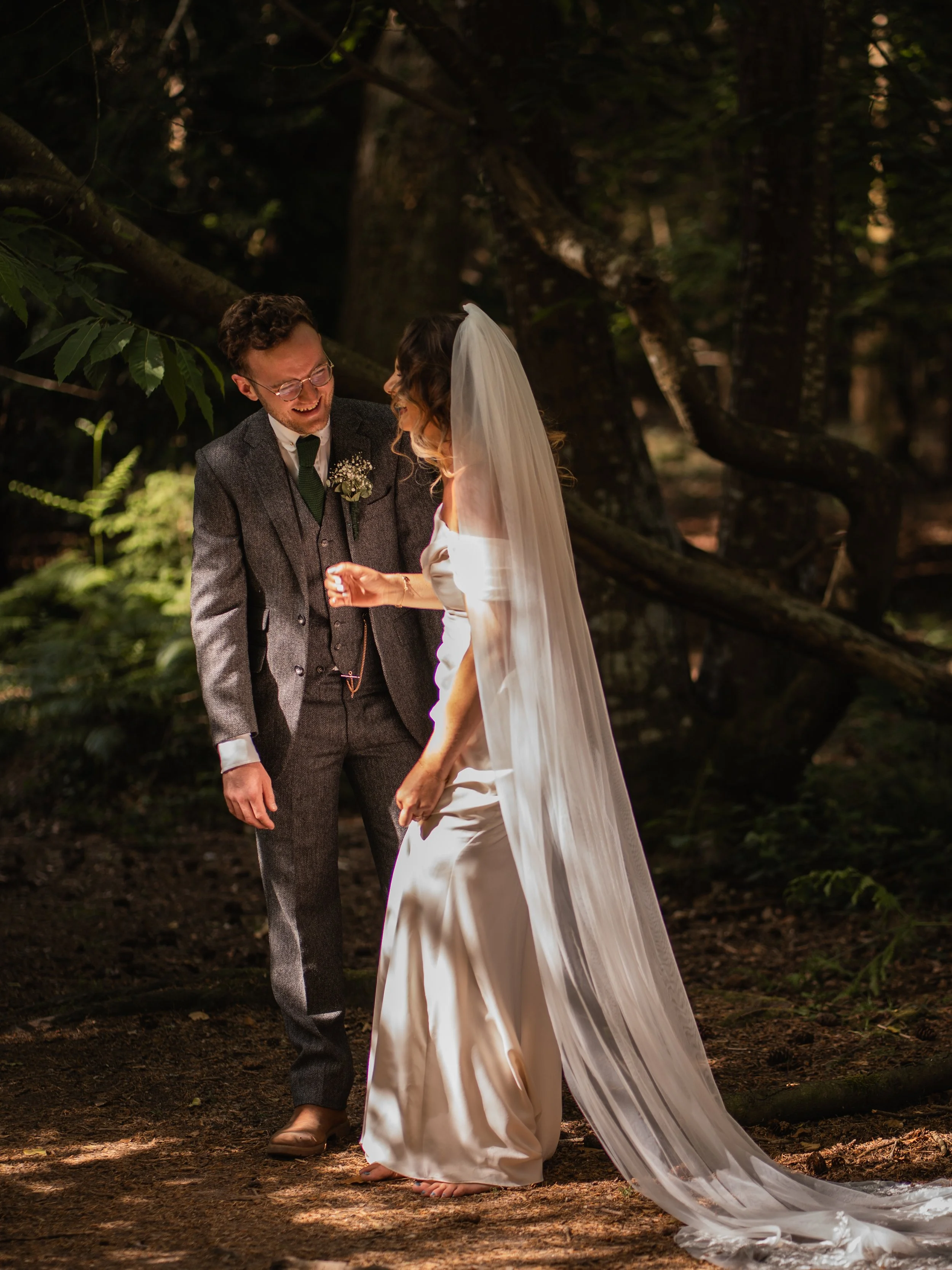 A bride and groom are laughing and holding hands in a forest with trees and foliage around them.