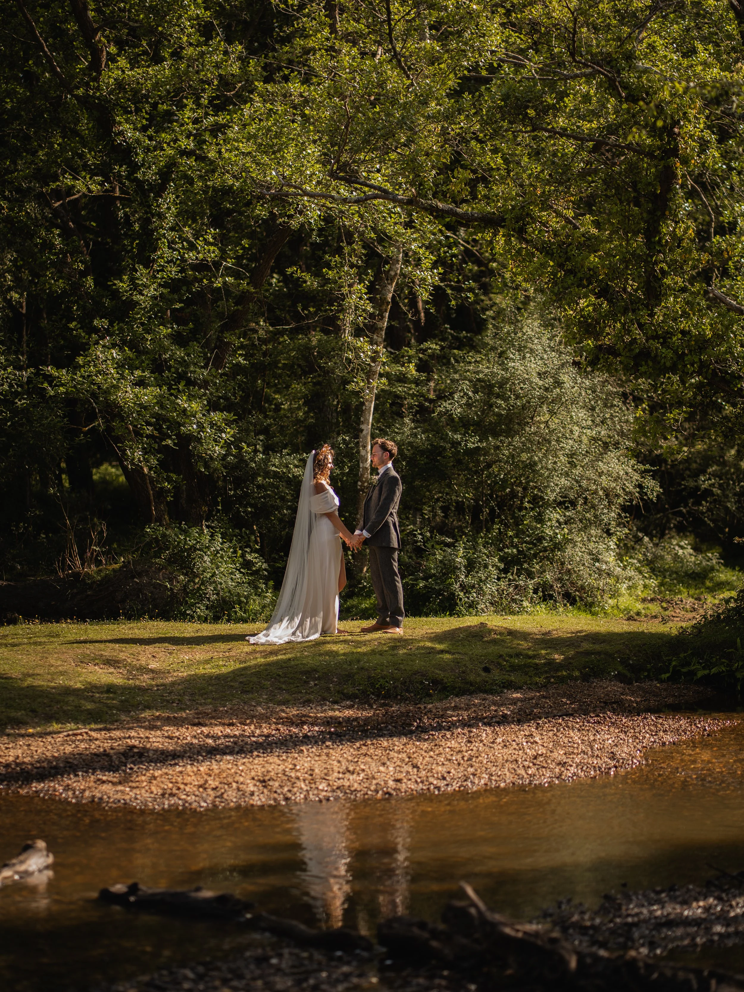 A bride and groom holding hands and facing each other outdoors near a body of water, surrounded by trees and greenery, during daylight hours.