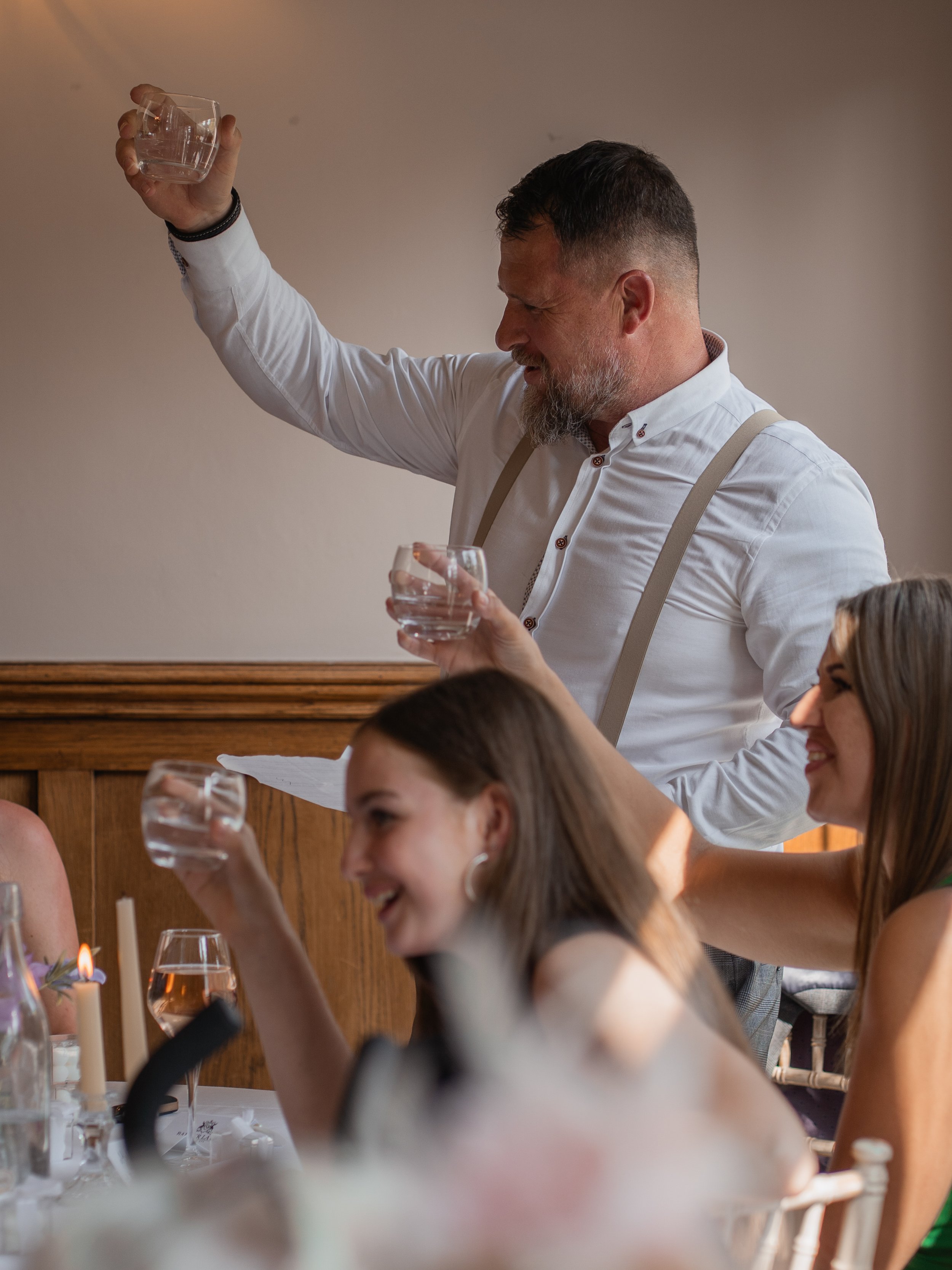 A man with a beard and a woman with long hair celebrating at a table, holding glasses of water, with a faint smile and joy on their faces.