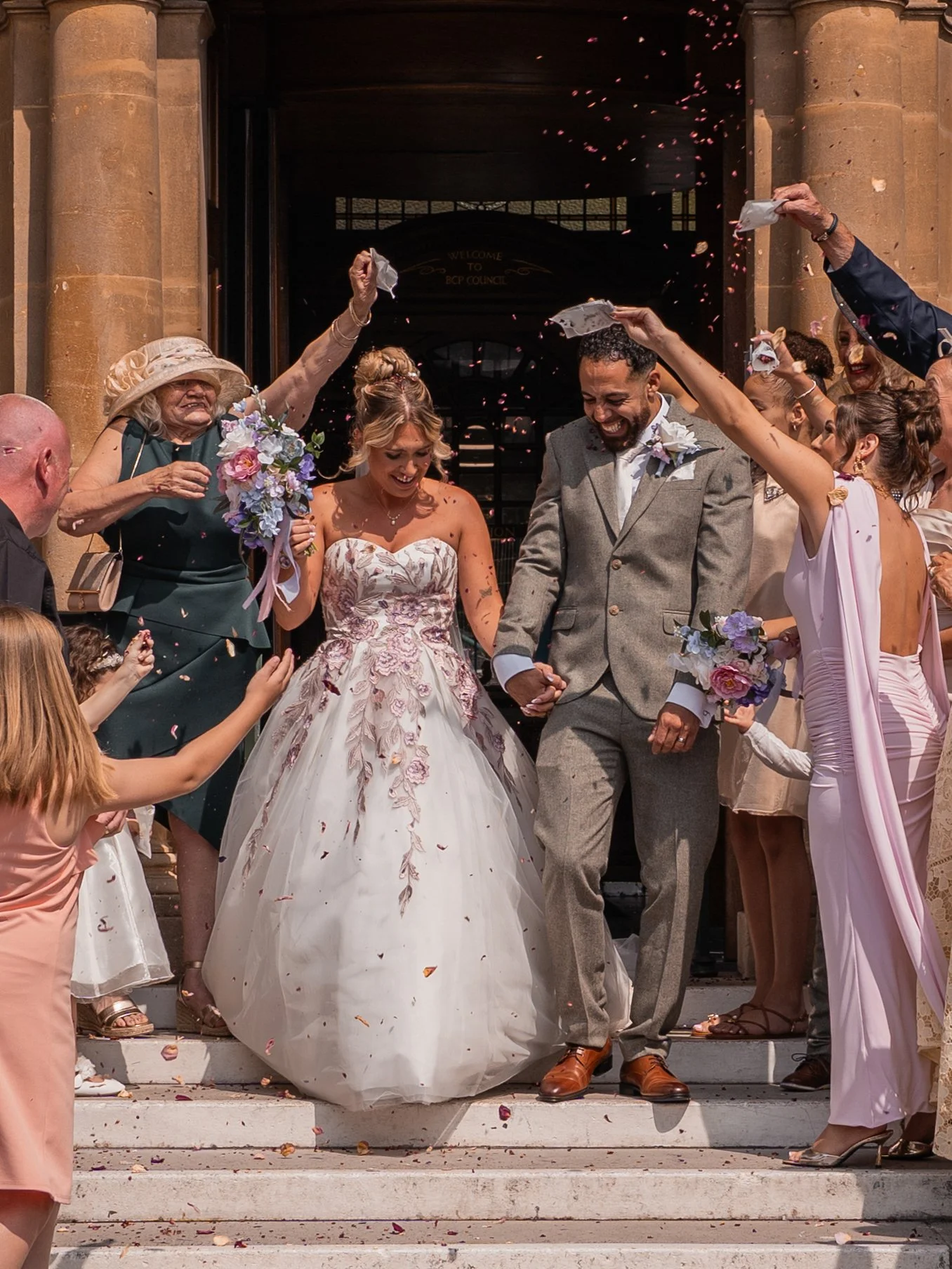 A newlywed couple leaving a church with friends and family celebrating their wedding outside the steps, confetti in the air, and the bride in a white ball gown and the groom in a gray suit.