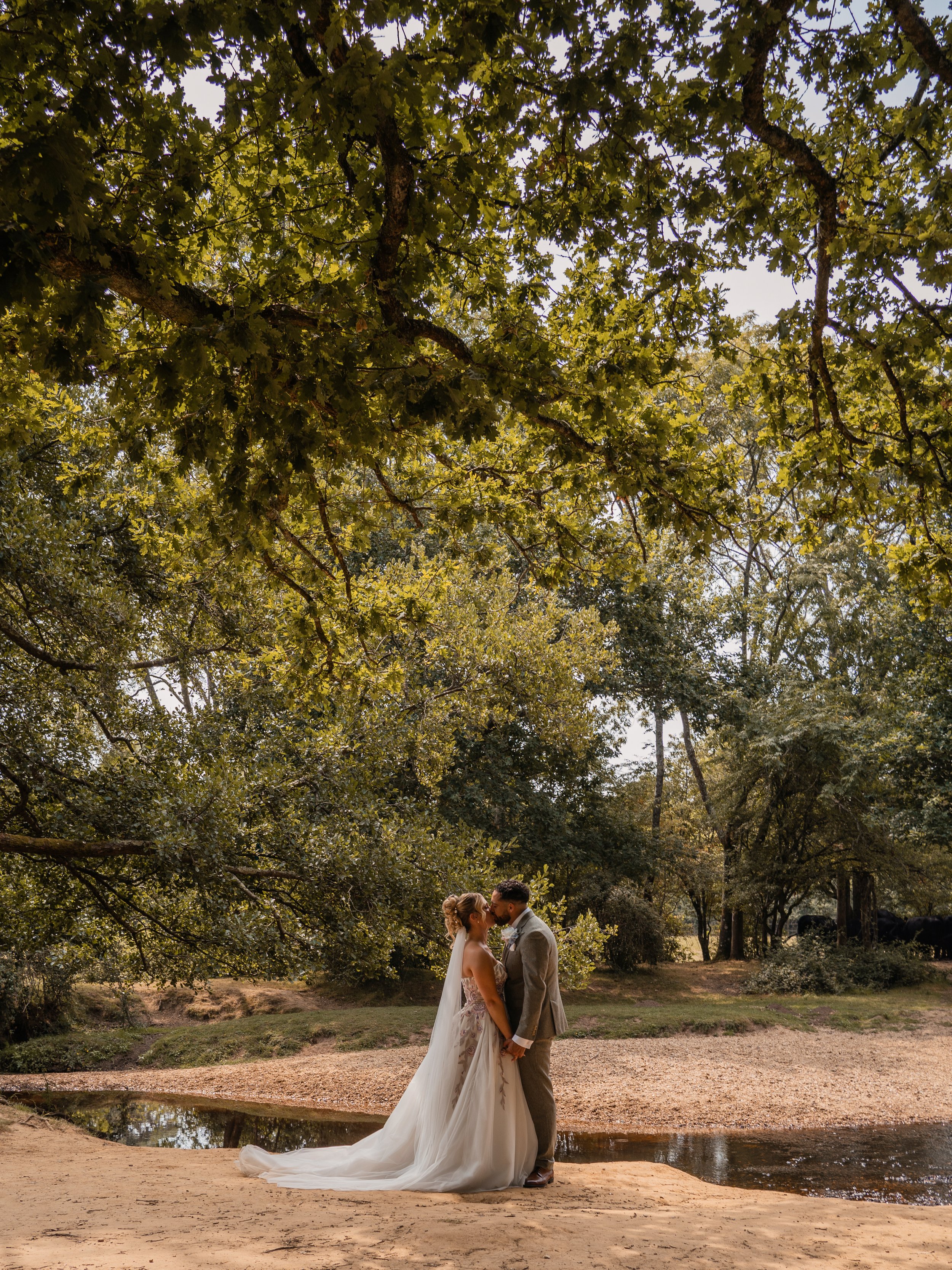 A bride and groom standing close together under large green trees in a park, with a small pond and a sandy area nearby, during their wedding ceremony.
