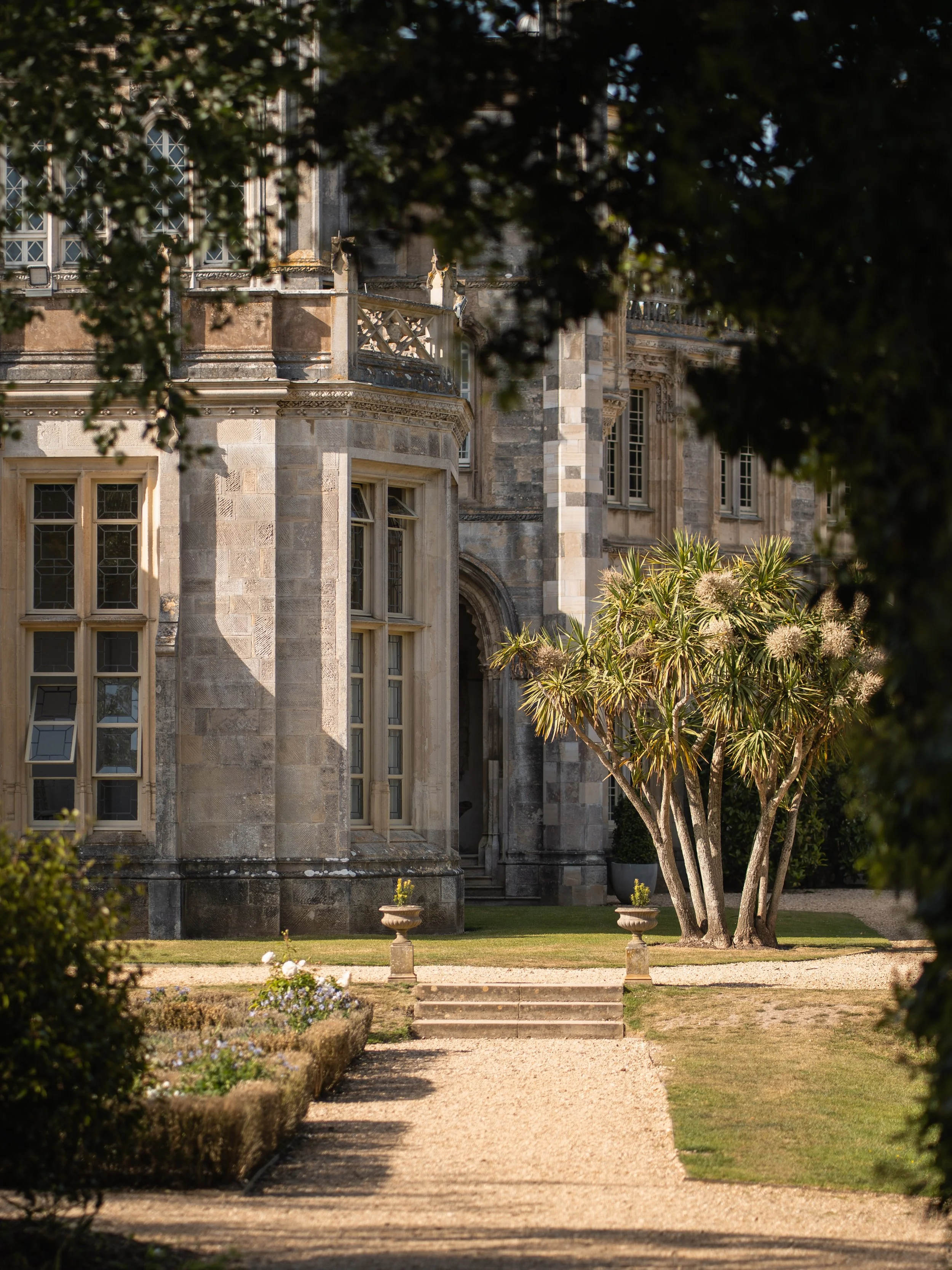 A historic stone building with large windows and ornate architectural details, framed by trees and a garden with a gravel pathway, potted plants, and manicured lawn.