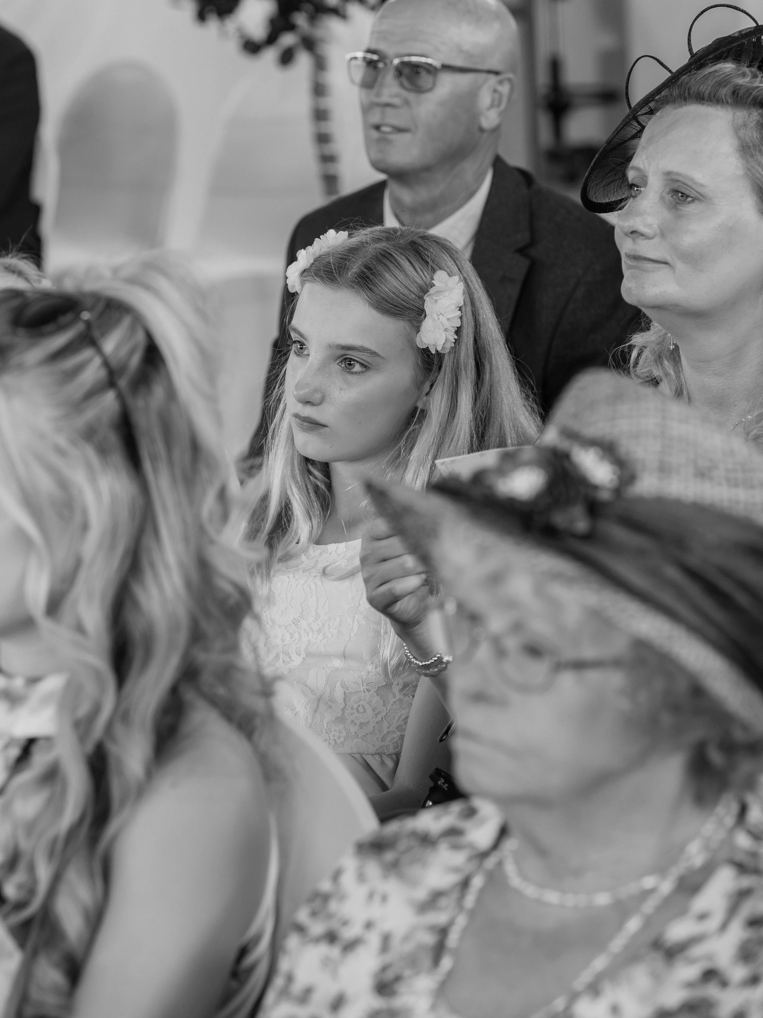 Black and white photograph of a group of people sitting together at an event, including a young woman with floral headband, an older man with glasses, and a woman wearing a fascinator hat.