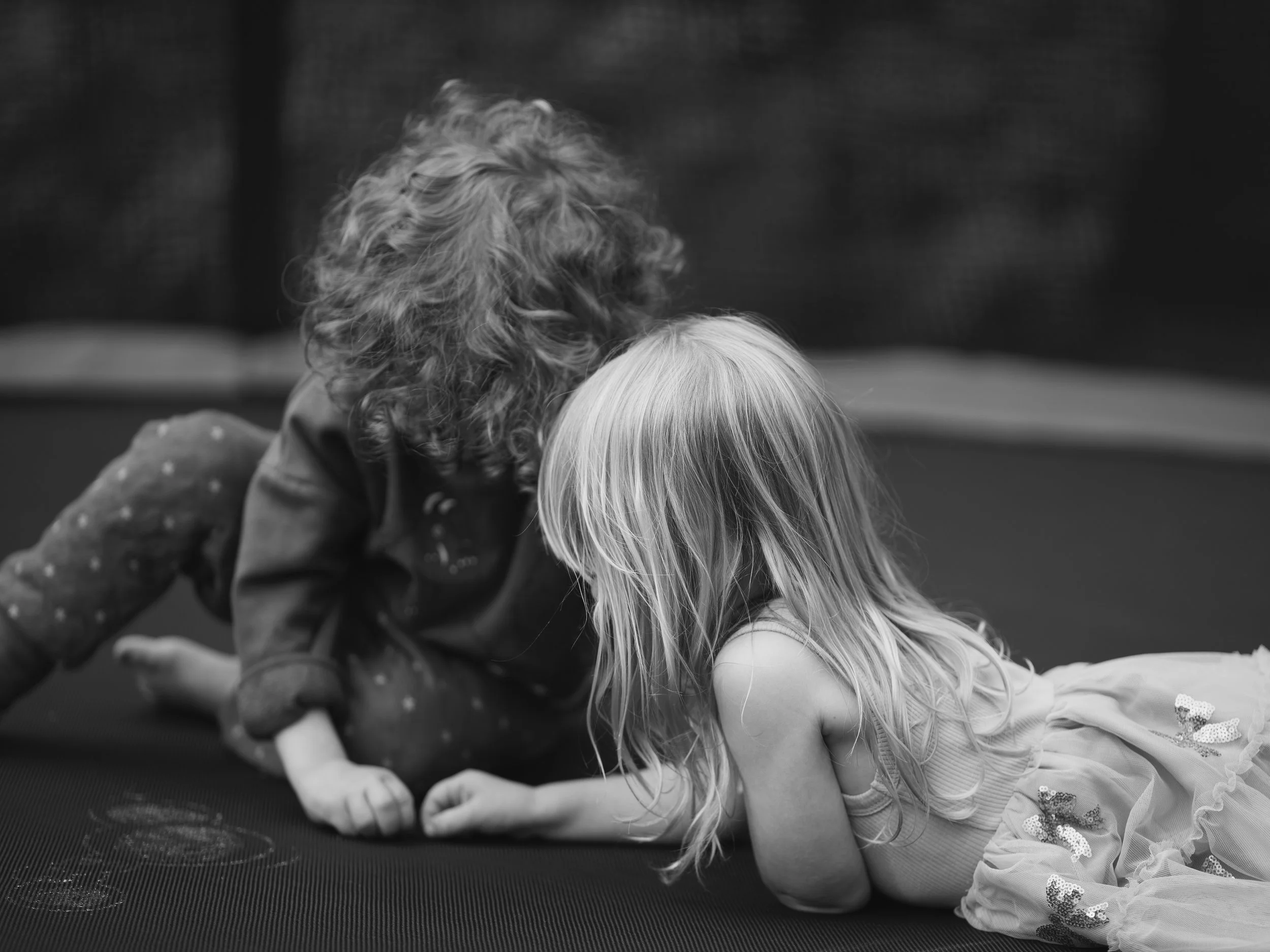 Black and white photo of a young girl with long hair in a costume, leaning forward on her hands, engaging with an adult woman with curly hair, both appearing to be playing or interacting on a dark surface.