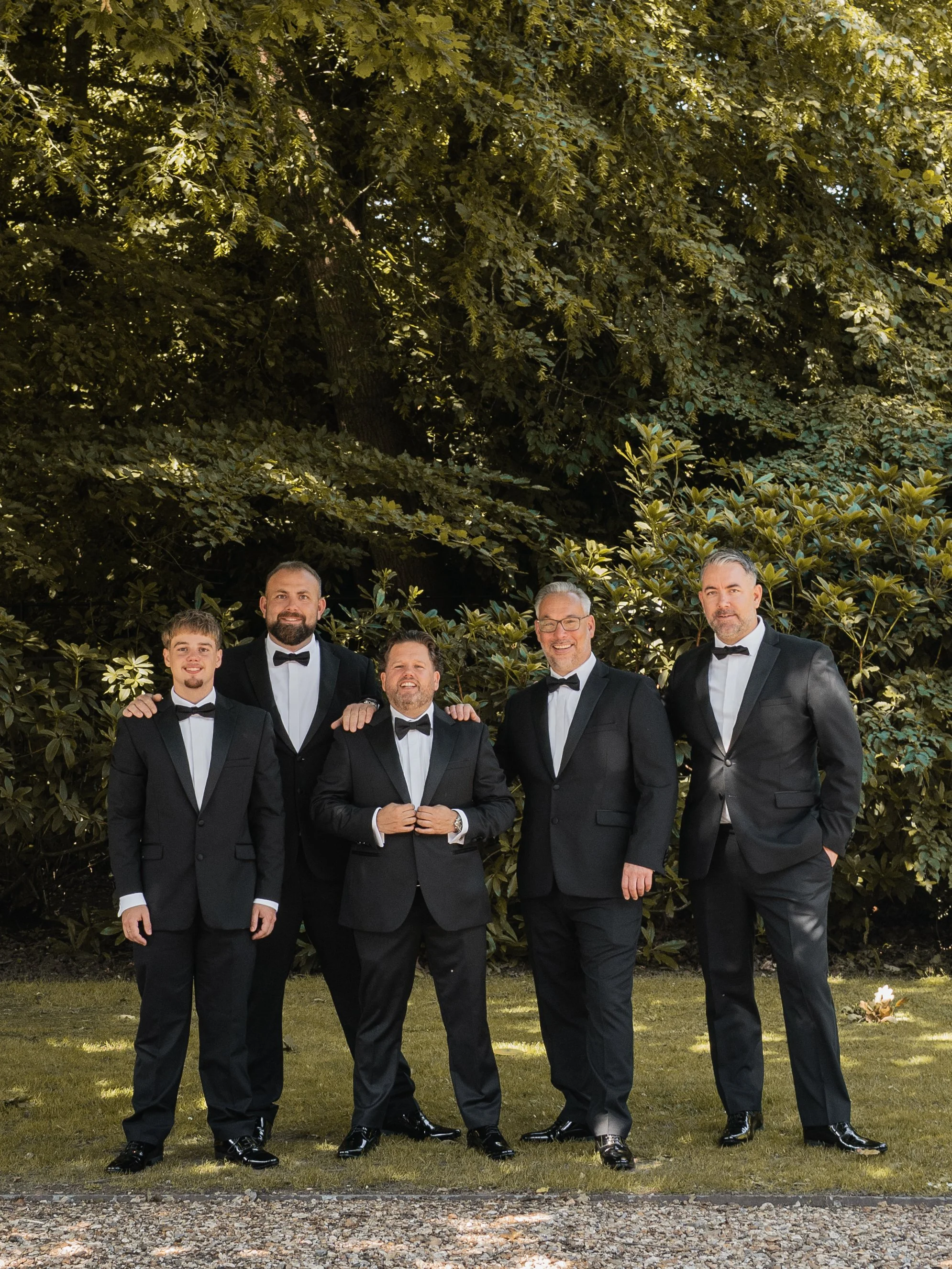 Group of six men in formal tuxedos with bow ties standing outdoors in front of lush green bushes and trees, smiling for a photo during daytime.