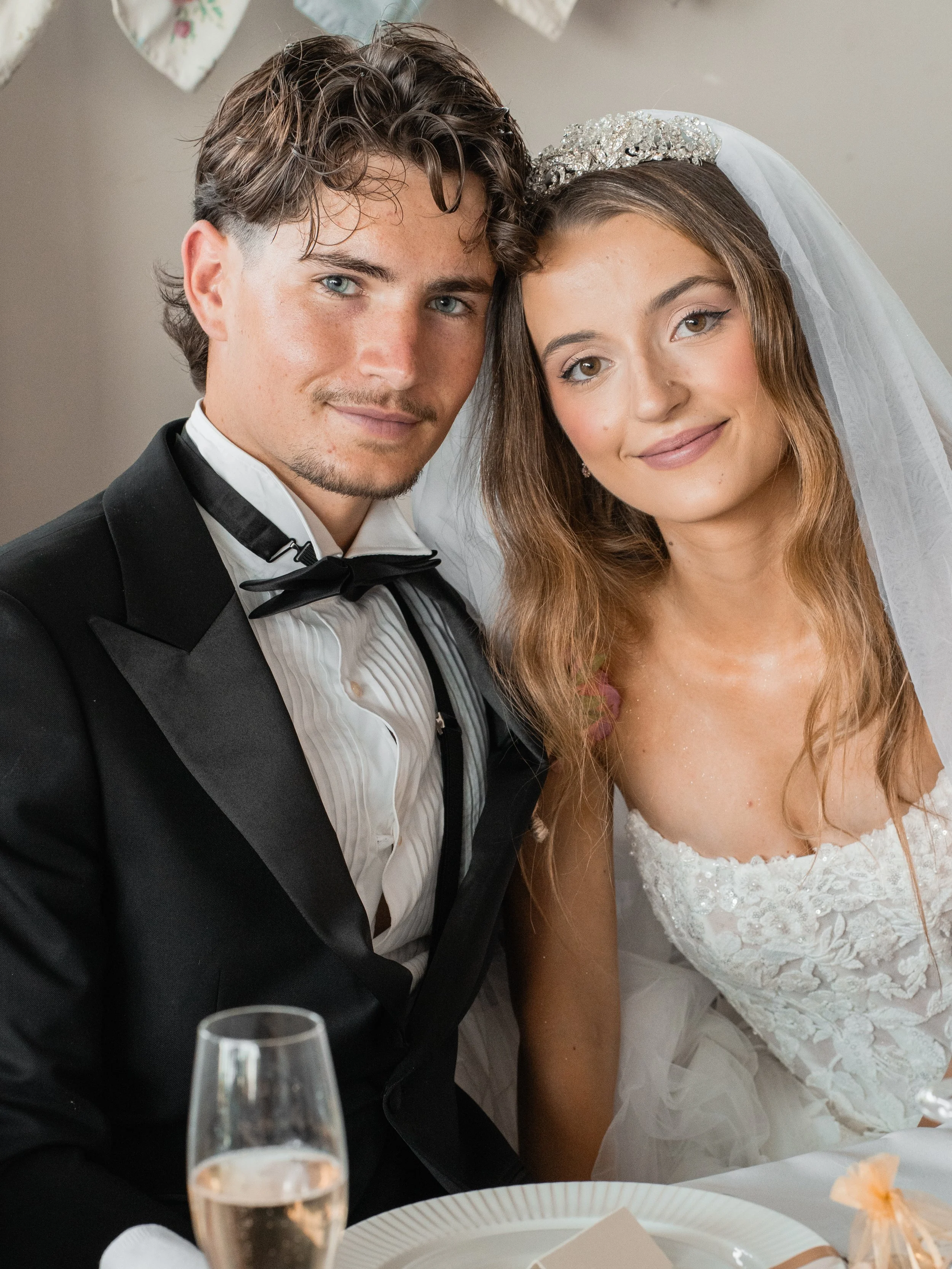 A young man and young woman dressed in formal wedding attire sitting close together at a celebration. The man wears a black tuxedo with a bowtie, and the woman wears a white wedding dress with lace details, a veil, and a decorative headpiece. They ar