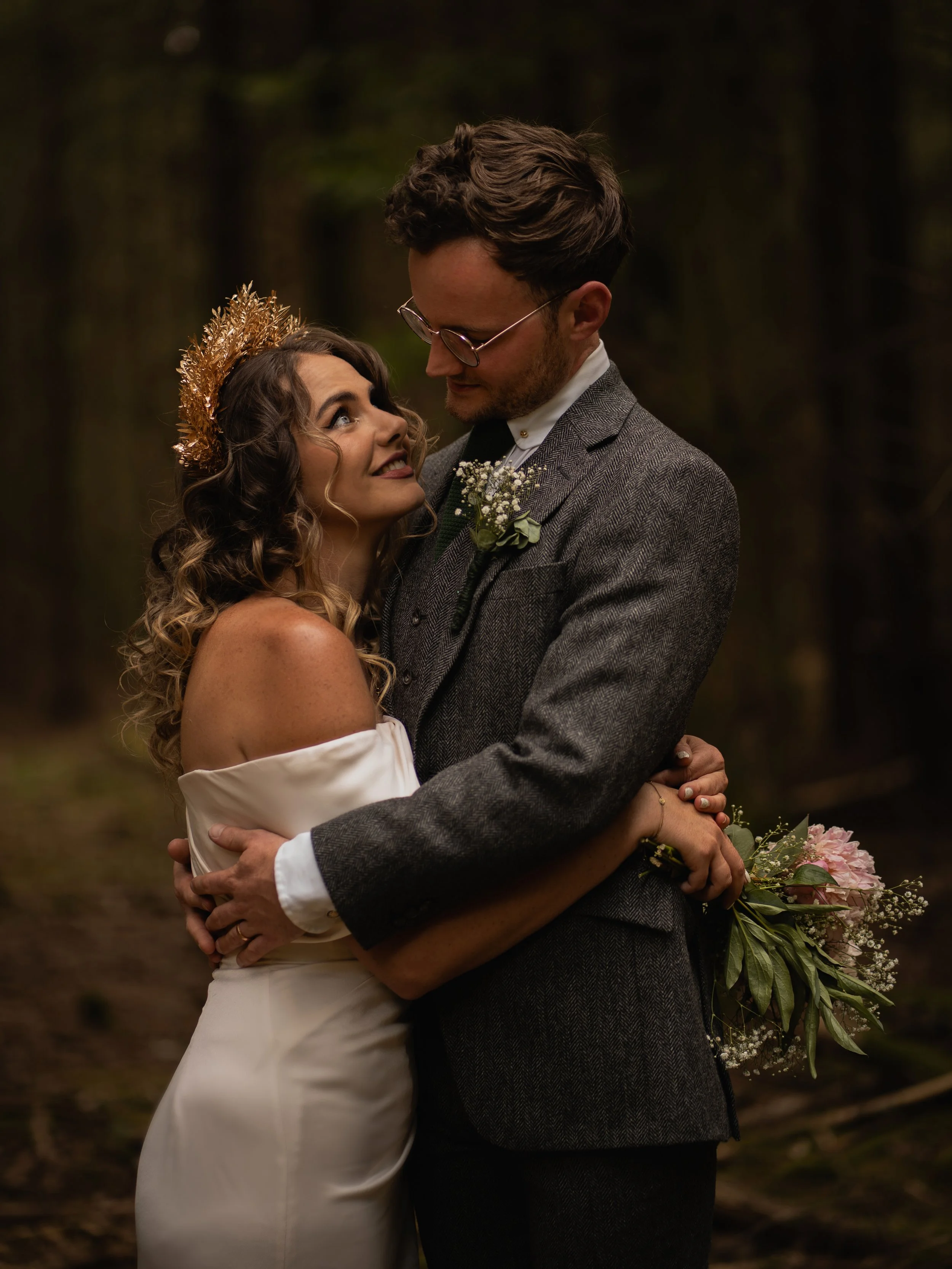 A bride and groom sharing a romantic embrace outdoors, with the bride looking up at the groom and both smiling gently. The bride has dark curly hair, a floral headpiece, and a white off-the-shoulder wedding dress. The groom has dark hair, glasses, a 
