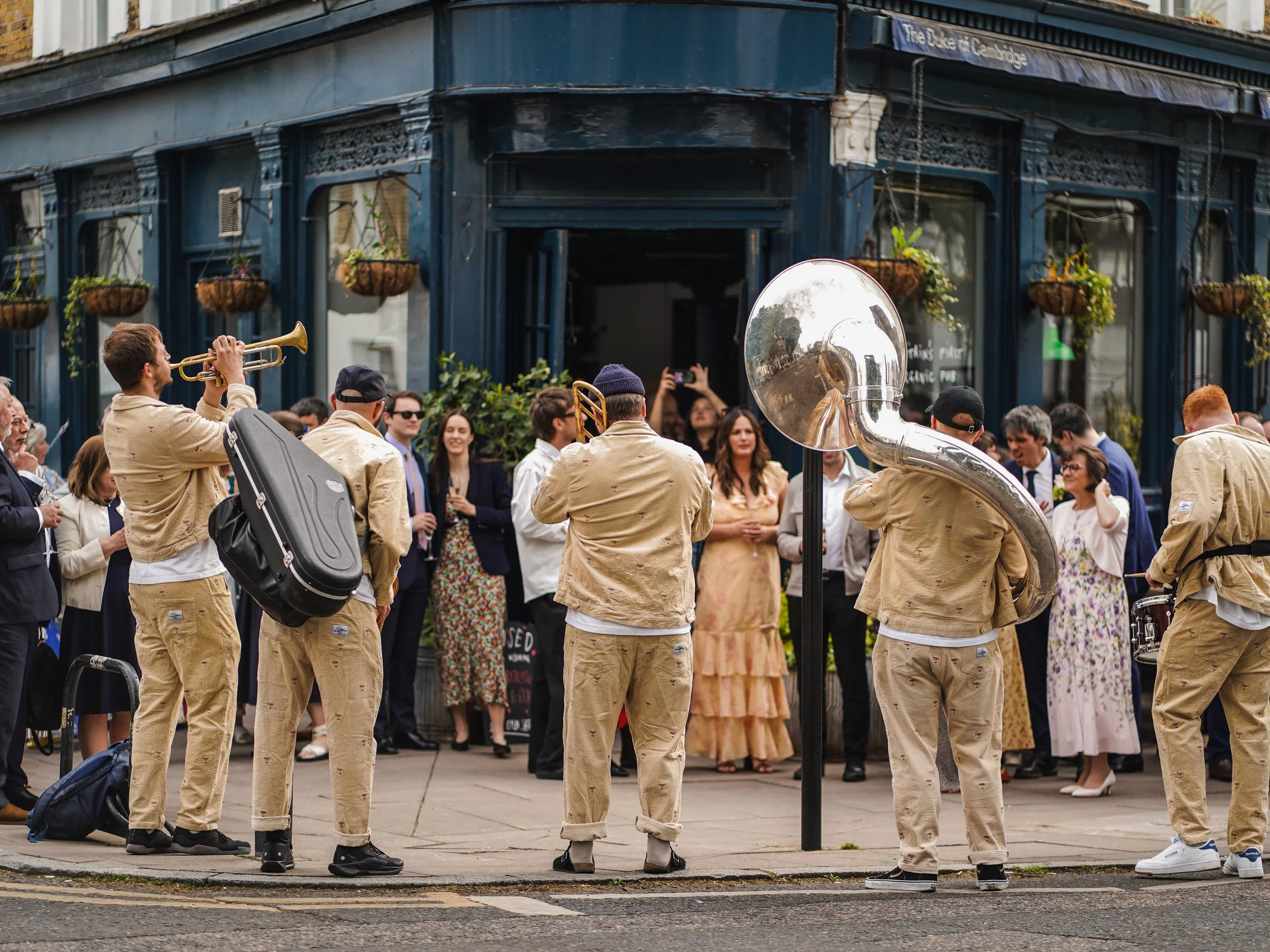 A street scene featuring a musical band playing instruments, with a crowd of onlookers in front of a blue building with hanging plants, during daytime.