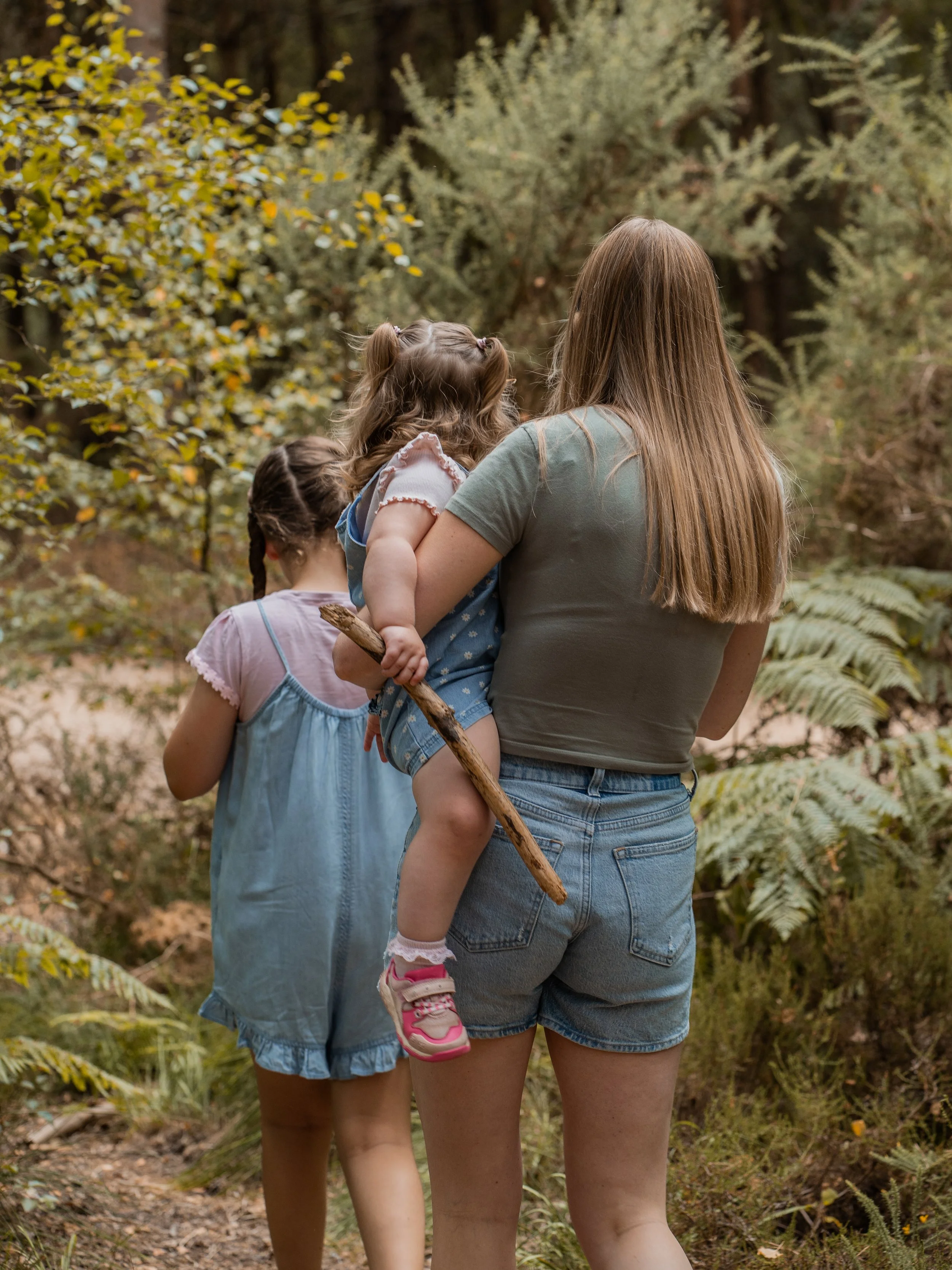 A woman carrying a young girl on her back through a wooded area, with another girl walking ahead. The scene is outdoors among trees and ferns.