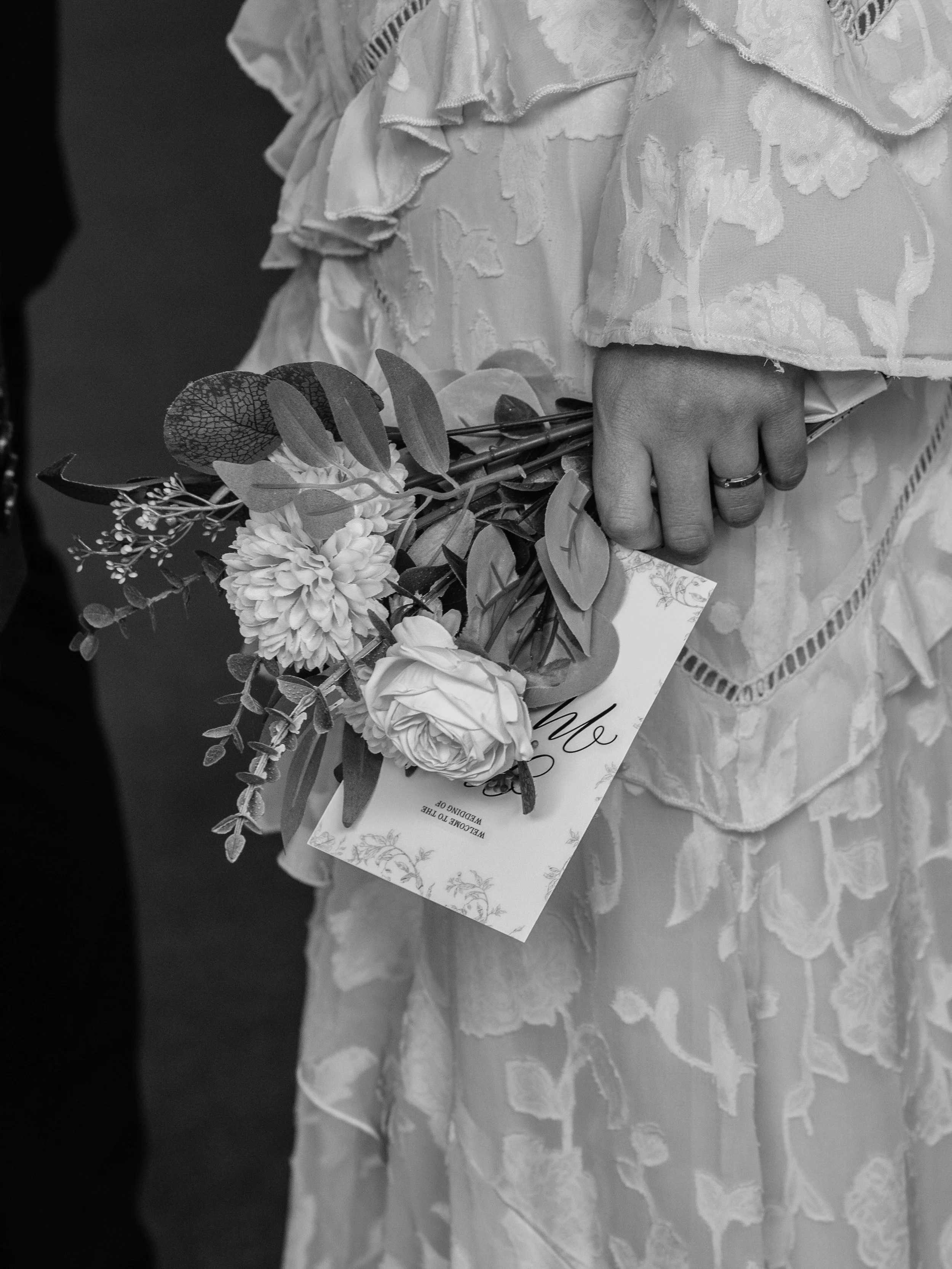 Black and white photo of a person in a lace dress holding a bouquet of flowers and a card.