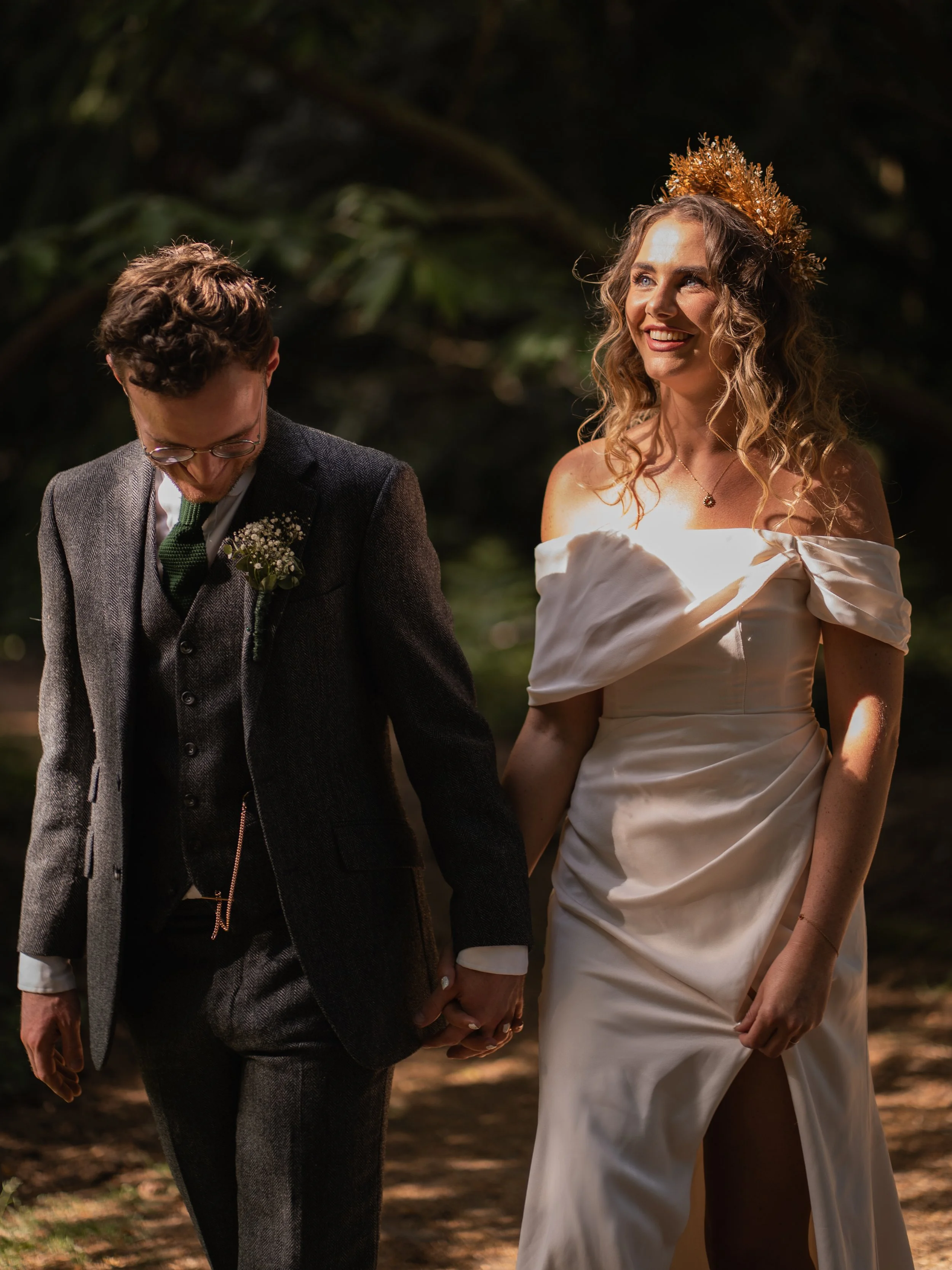 A smiling woman wearing a white off-shoulder dress and a gold crown, holding hands with a man dressed in a gray suit with a boutonniere, in a dark, outdoor natural setting.