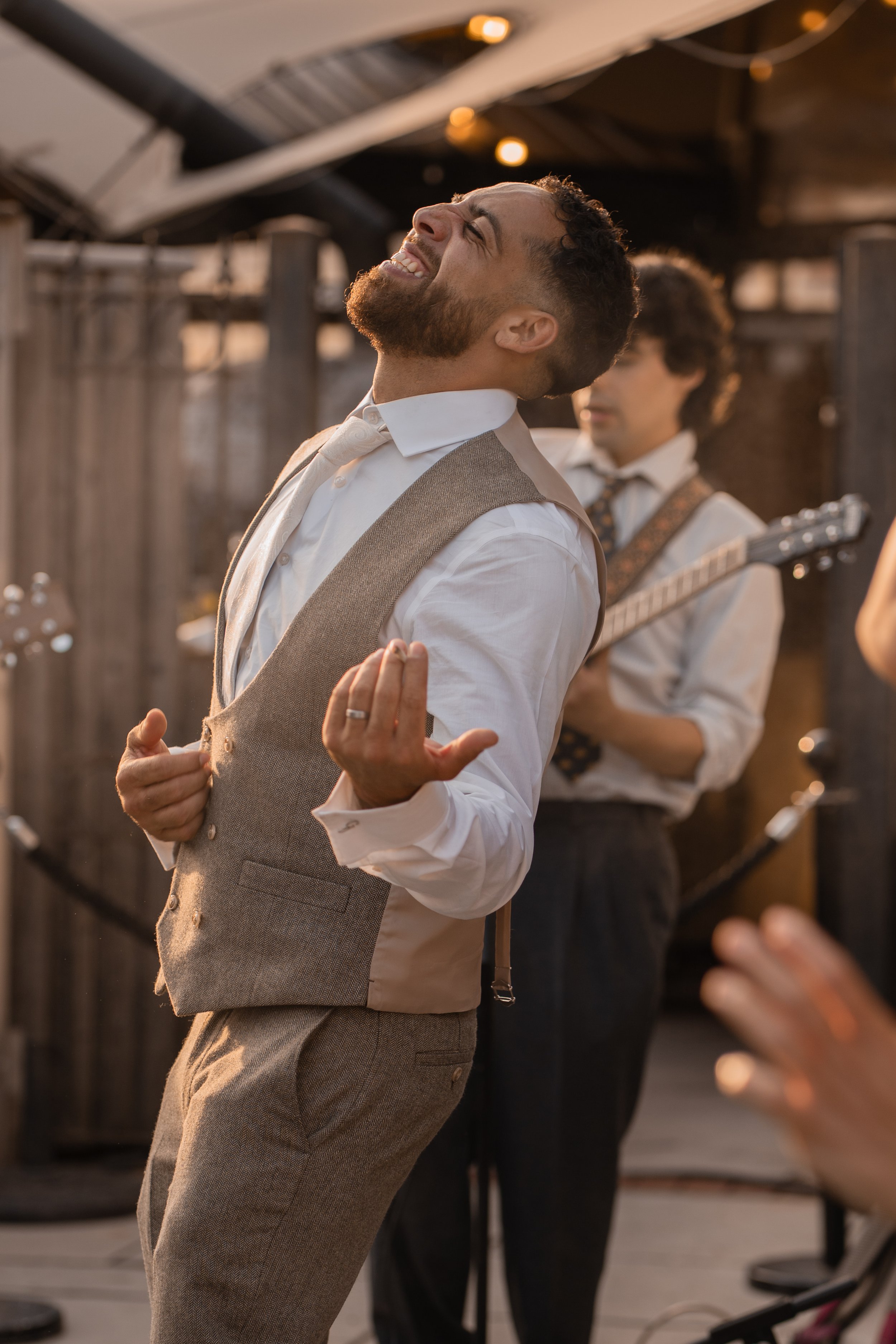 A man with a beard and curly hair is wearing a white shirt and a beige vest, looking upwards with an expression of joy, while dancing outdoors during sunset. Another man in the background holds a guitar, also dressed in a white shirt with suspenders,