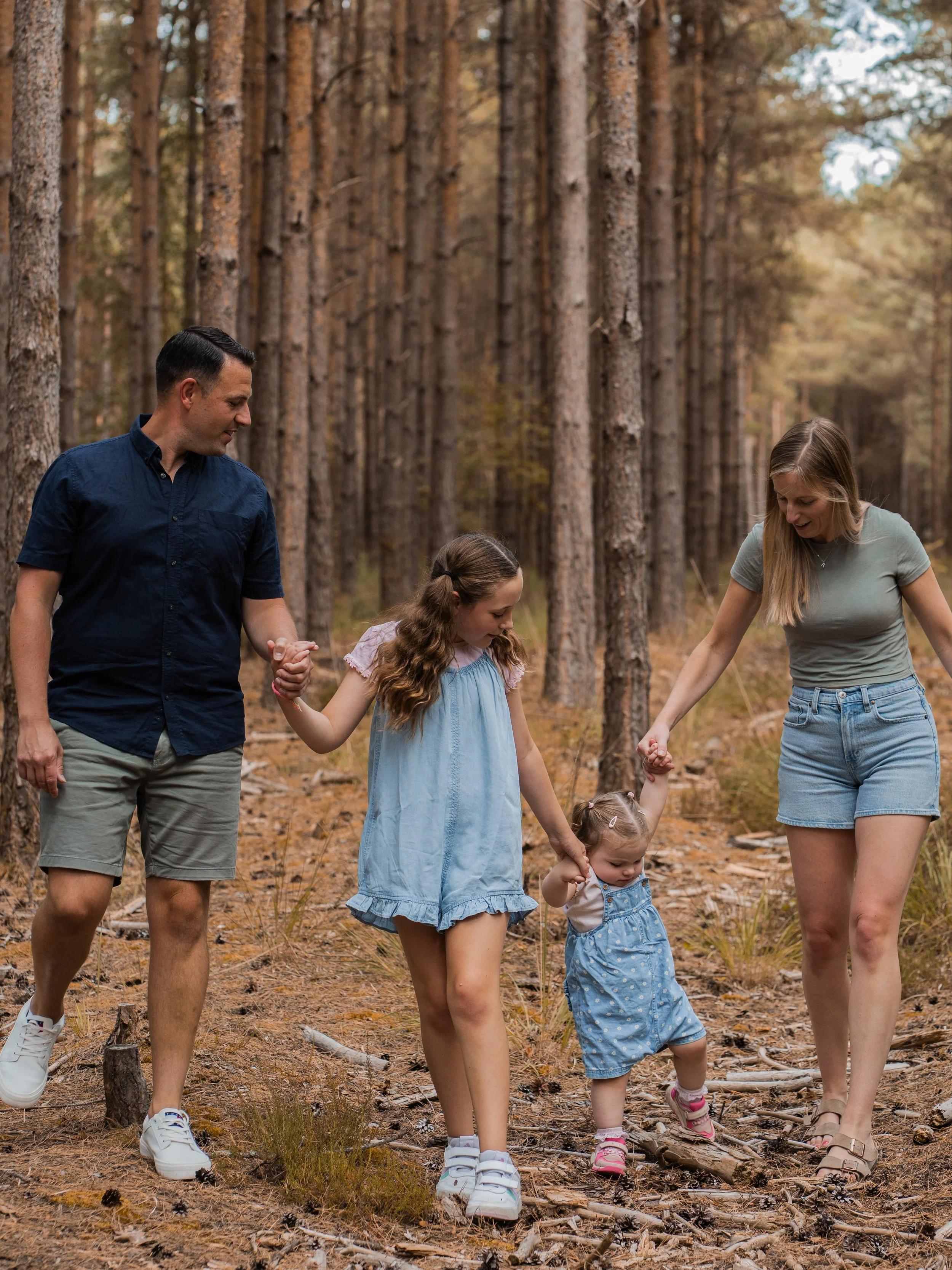 A family of four walking hand in hand through a forest trail during autumn, among tall pine trees.