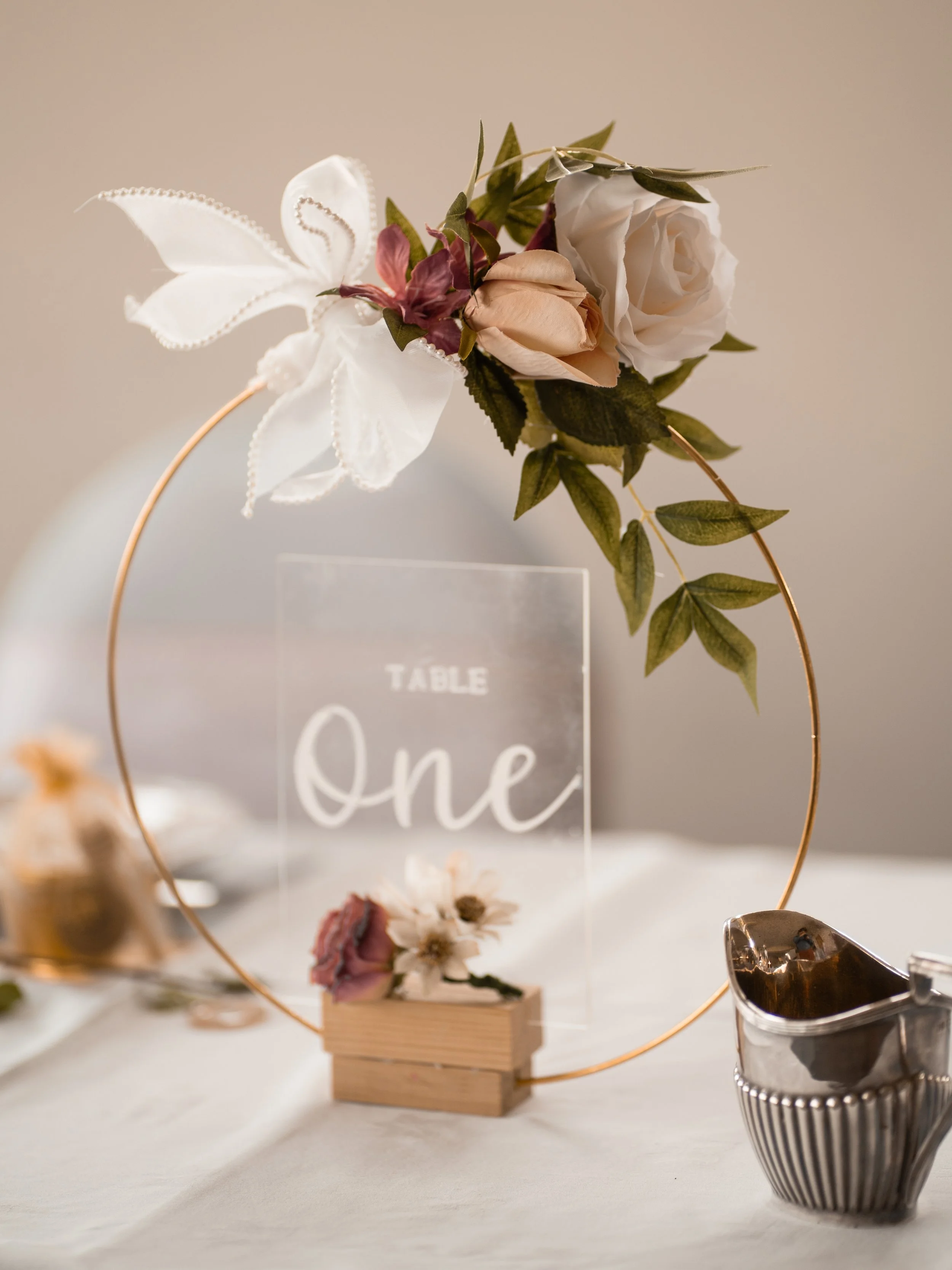 Elegant table centerpiece with flowers, a glass sign reading 'One,' and decorative items, set on a white table.