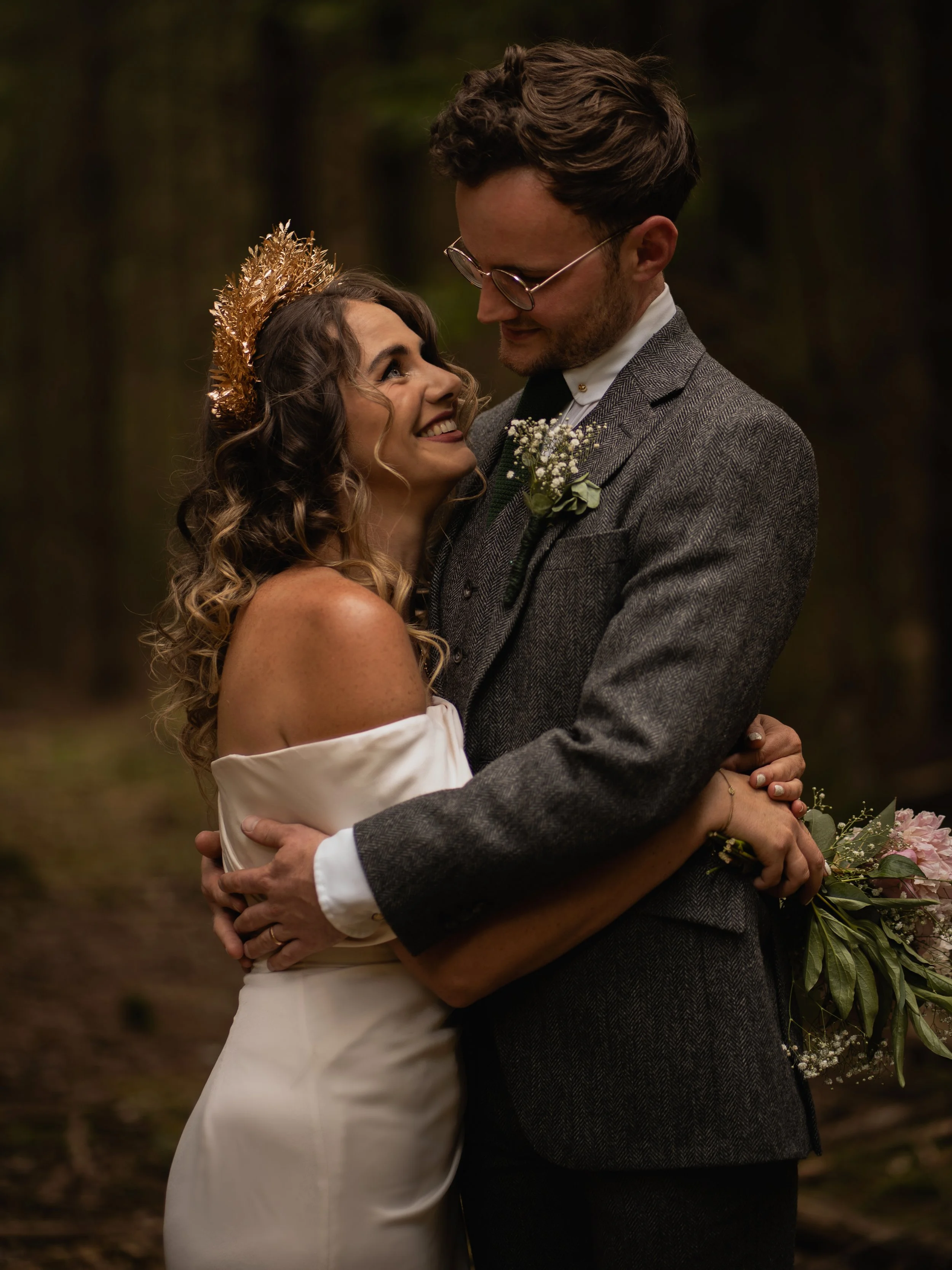 A couple in wedding attire embracing outdoors, with the woman smiling at the man, who is looking down at her. The woman wears a white off-shoulder dress and a gold crown, and the man wears glasses, a grey tweed suit, and a boutonniere. The background