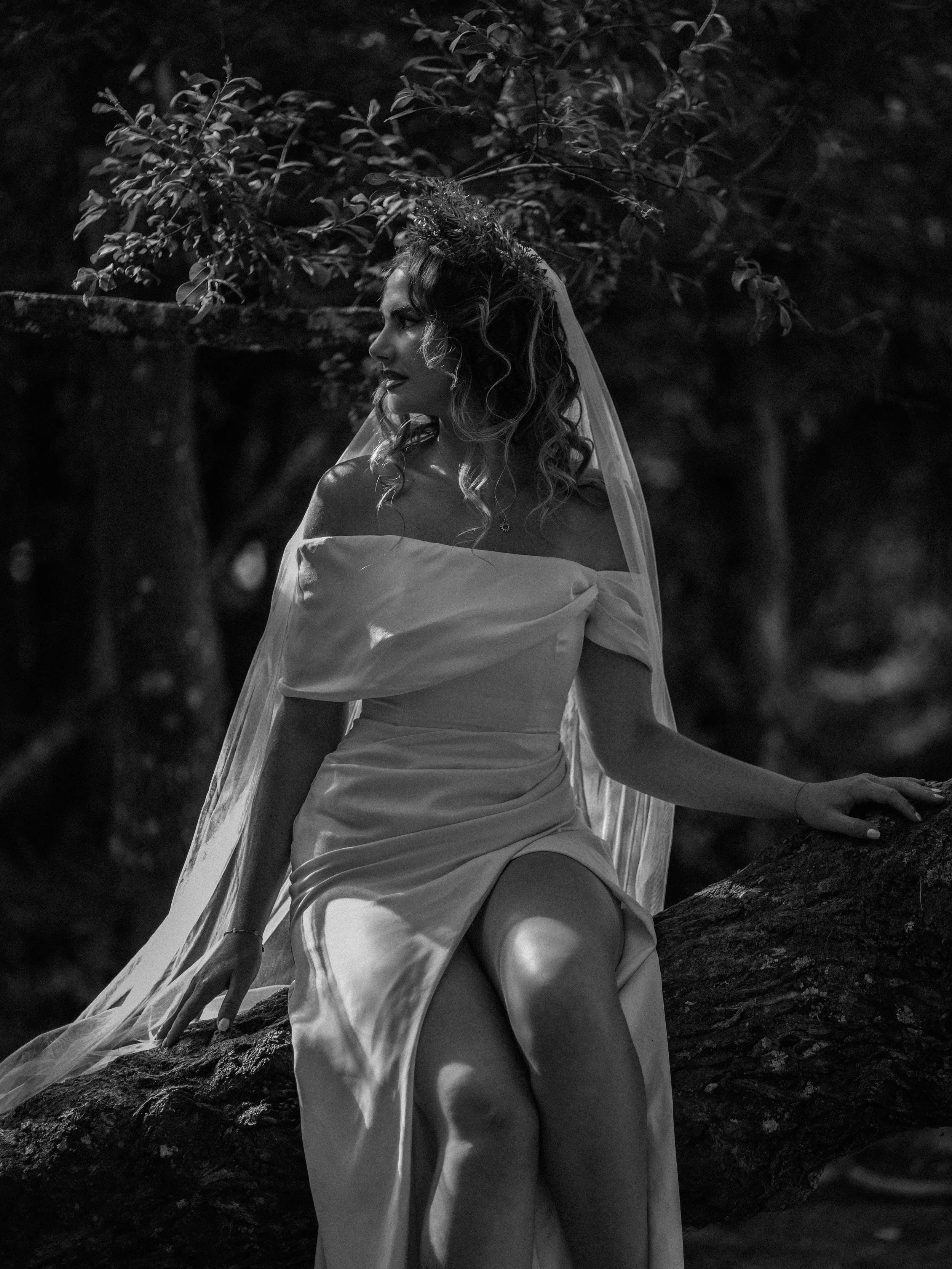 A woman with curly hair wearing a wedding dress and veil, sitting on a tree branch outdoors at night.