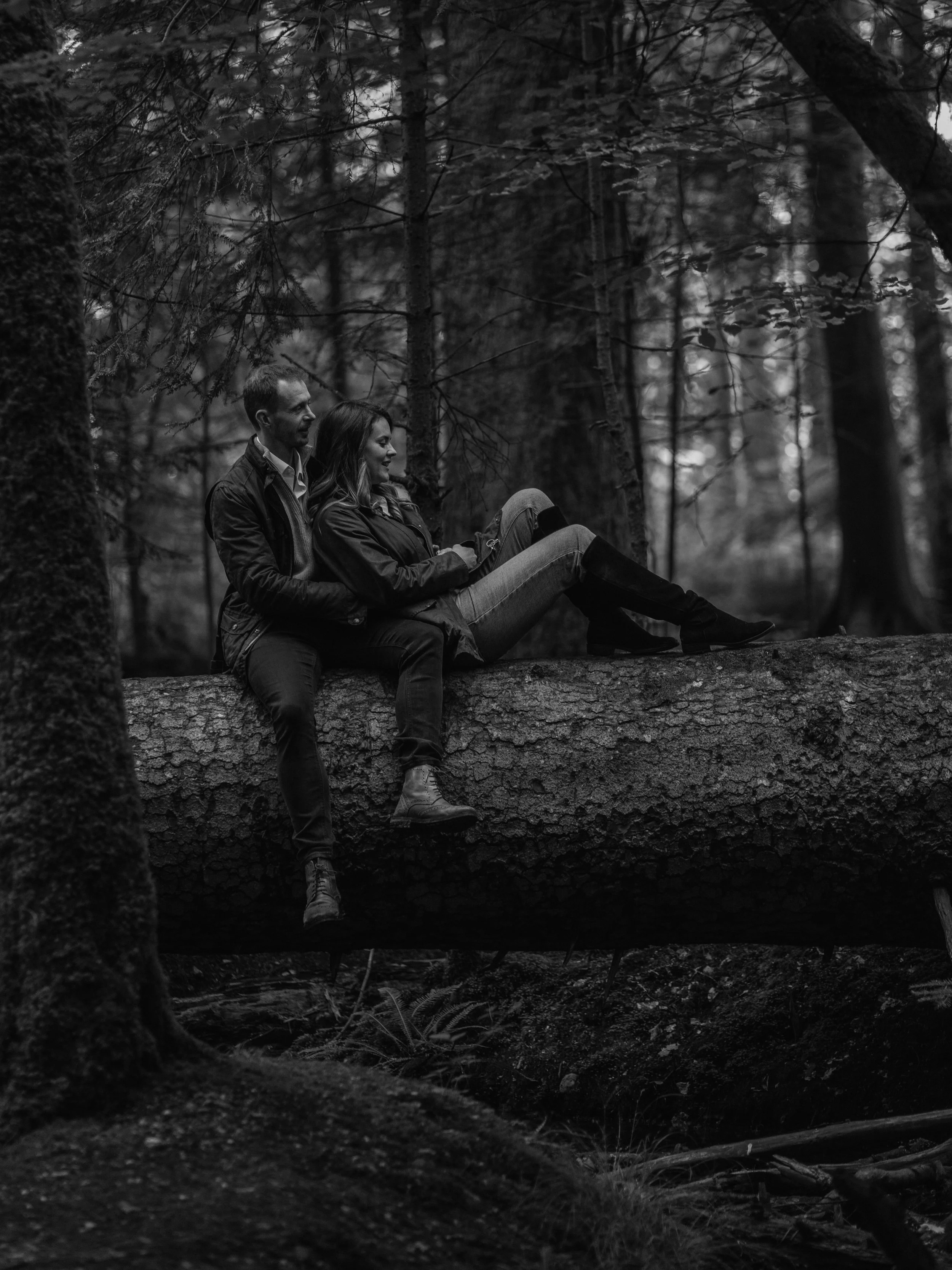 A black and white photo of a man and woman sitting on a large fallen tree trunk in a dense forest, with trees and foliage surrounding them.